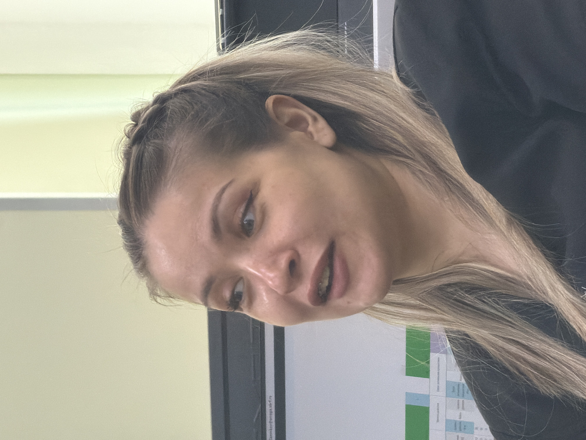 A fair-skinned young woman with blonde hair and a braided crown is the central figure. She is positioned in front of a whiteboard containing charts and text in Russian. She is wearing a dark-colored garment. The location is identifiable via text on the whiteboard as Krasnoyarsk, Russia.