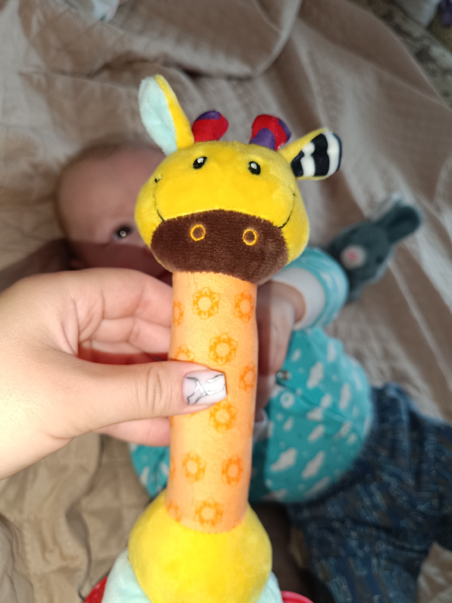 The image shows a person's hand holding a plush giraffe toy in front of a baby lying on a brown textured surface, possibly a bed. The giraffe toy has a yellow head, a brown muzzle, and an orange neck with a floral pattern. The baby is wearing a blue and white patterned outfit and has a gray bunny toy nearby. The location is Tobolsk, Russia.