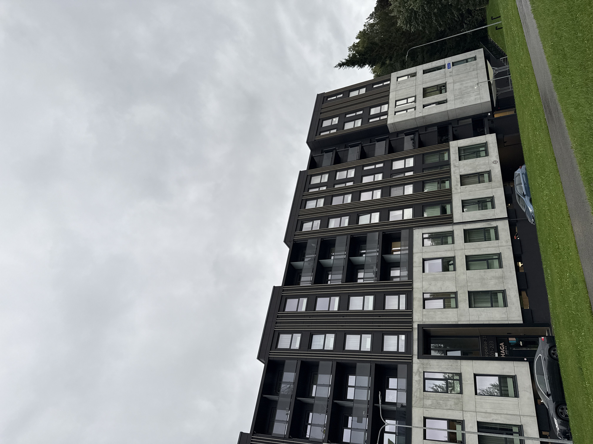 The image captures a modern apartment building with a distinctive angled facade, located in the Mustamäe district of Tallinn, Estonia. The building is primarily black with grey concrete elements, featuring multiple stories with numerous windows and balconies. The sky above is overcast and grey. In the foreground, a sloped grassy area with a paved path runs alongside the building. Two cars are parked on the street in front of the building, one dark grey sedan and one blue hatchback. The entrance to the building is visible, with signage indicating "EPESEELSI 31" and "HAGA HOME". Trees are visible behind the building to the right, suggesting a somewhat suburban or semi-urban setting. The overall scene depicts an urban residential environment on a cloudy day.