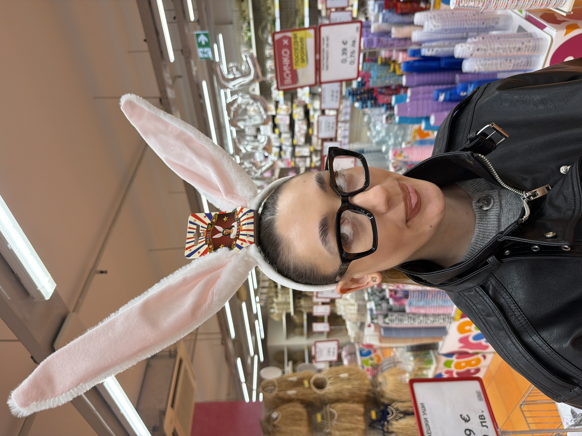 A woman wearing bunny ears and black-framed glasses stands in a store, likely in Sofia, Bulgaria. She is wearing a black leather jacket over a gray sweater. The store appears to sell a variety of party supplies and decorations, as indicated by the shelves filled with items in the background. Price tags in Bulgarian currency (lev) are visible on the shelves. The lighting is provided by fluorescent ceiling lights.