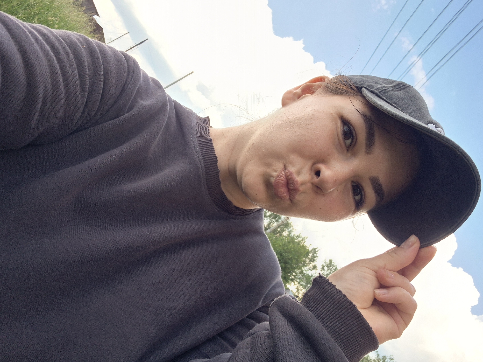 A young woman wearing a dark grey baseball cap and a dark grey sweatshirt is taking a selfie outdoors. She is making a kissing face with her lips puckered. She is holding the brim of her cap with her right hand. The background consists of a bright blue sky with fluffy white clouds, some trees, and green grass. In the distance, there appears to be a building and some electrical wires. The lighting suggests it is daytime. The image is taken from a low angle, looking up at the woman.