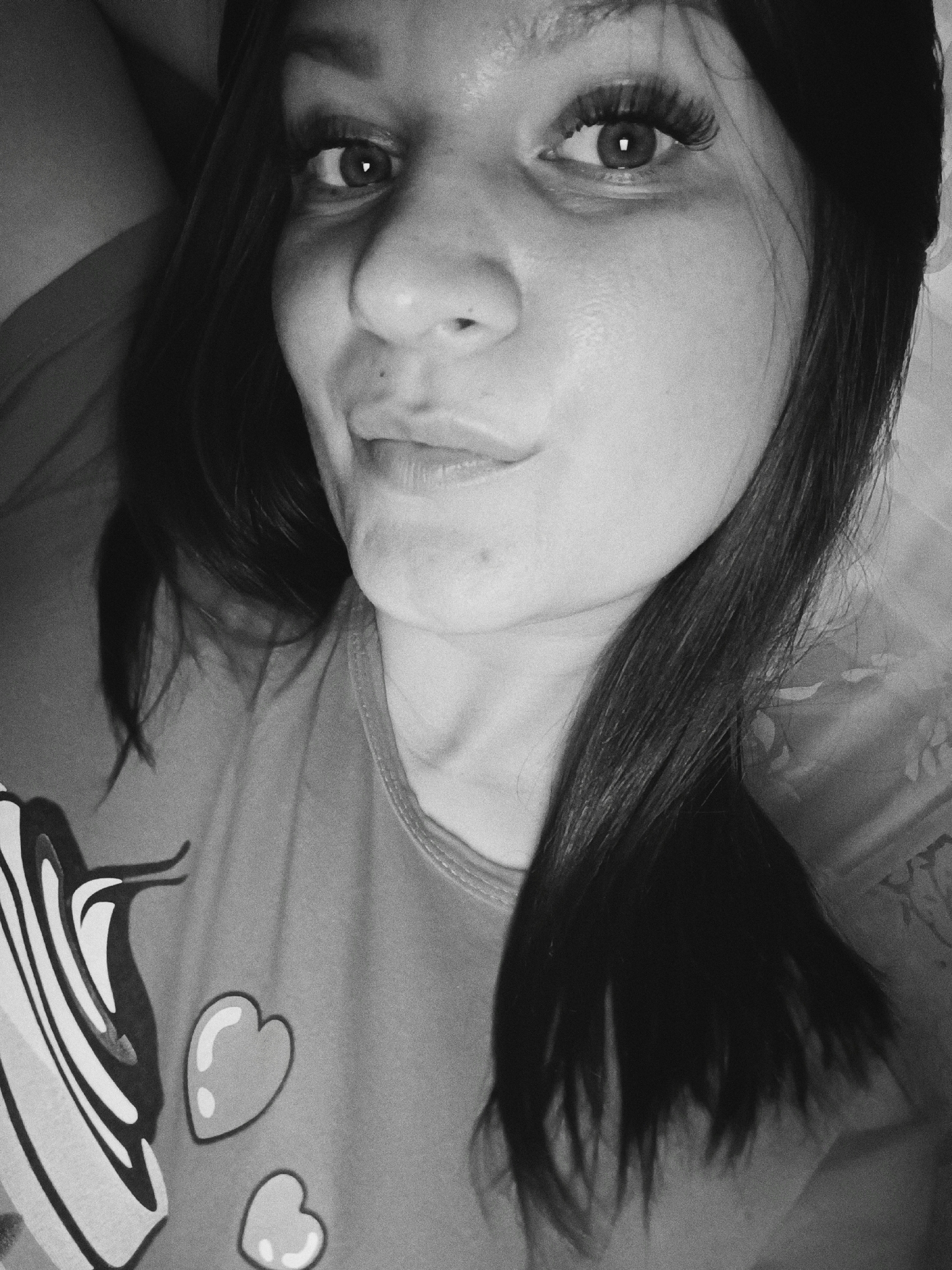 This black and white image presents a close-up, high-angle portrait of a young adult female individual, positioned indoors within a private residence in the city of Tobolsk, Russia. The subject has long, dark hair parted to one side, which falls across her shoulders. She gazes directly at the camera with her eyes open, exhibiting prominent eyelashes. Her lips are slightly pursed in a gentle expression. Visible on her face are minor skin imperfections, including small moles or blemishes. She is clad in a light-toned t-shirt or sleepwear top featuring a graphic design on the left side of her chest. This design includes a large, vertically striped object resembling a pastry, accompanied by two smaller, outlined heart shapes below it. The individual appears to be reclining, with a darker, plain surface partially visible behind her to the left, and a lighter, patterned fabric or material to her right. The image exhibits a grainy texture consistent with low-light conditions or specific photographic processing.