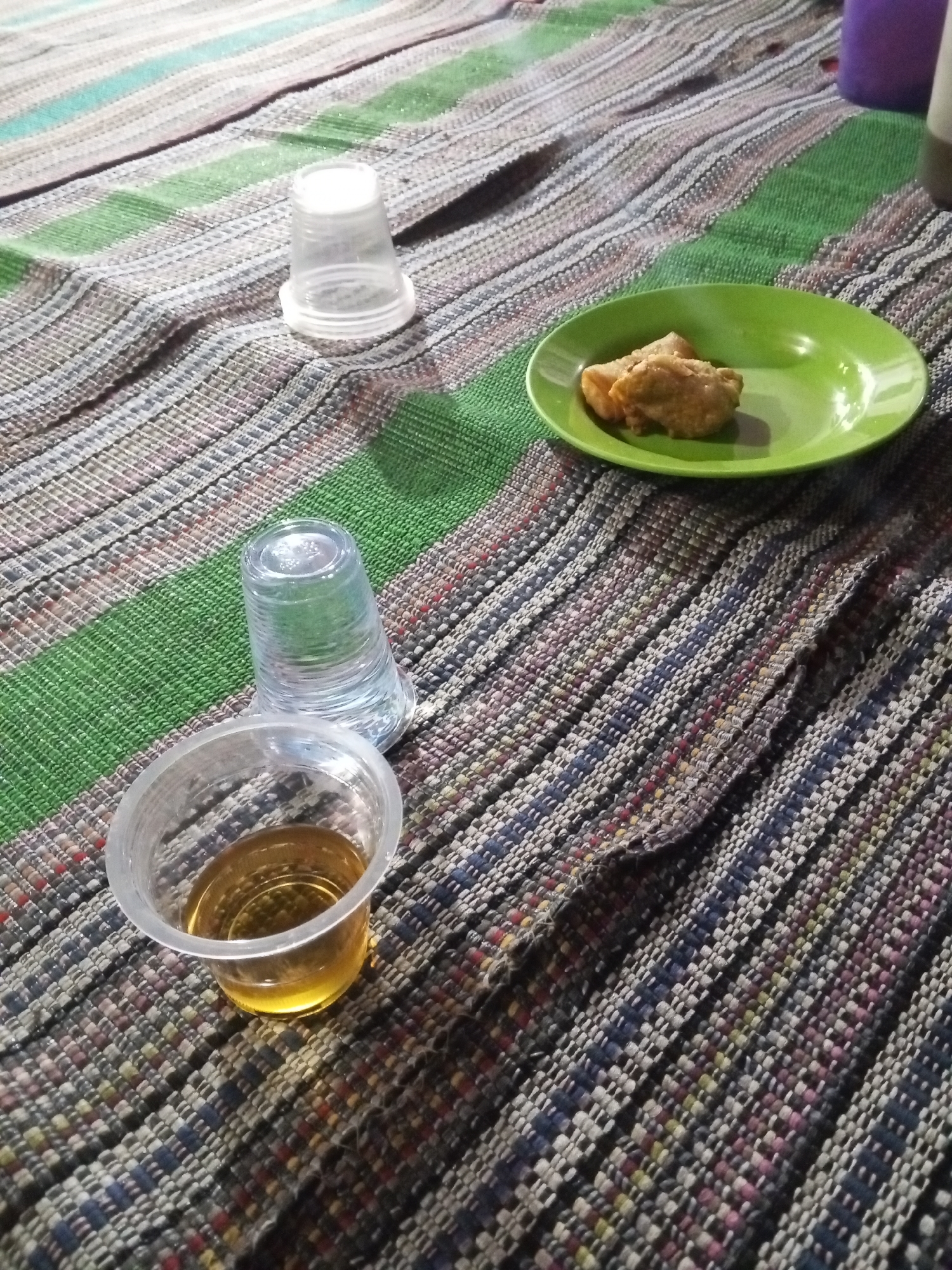 The image depicts a casual outdoor setting, possibly a street-side food stall or a home yard, in Ciruas, Indonesia. The focal point is a woven mat with a striped pattern of green, white, and brown hues, laid out on a surface. On the mat are three plastic cups and a small green plate. One cup, positioned in the foreground, contains a golden-brown liquid, likely tea or coffee. Adjacent to it is an empty, clear plastic cup. To the right of these cups, on the green plate, are two pieces of fried food, possibly tofu or a similar snack. Another empty, clear plastic cup is situated further back on the mat, near a blurred background element that appears to be a larger container, possibly purple. The lighting suggests it is evening or nighttime, with artificial light sources illuminating the scene. The overall impression is one of a simple meal or snack being enjoyed.
