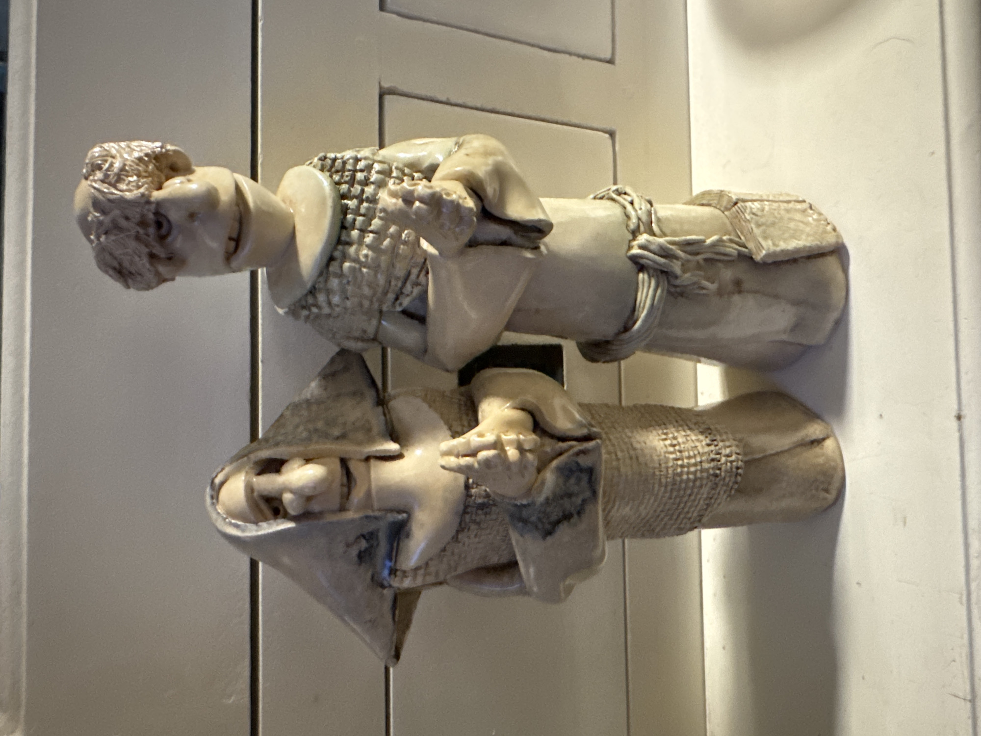 Two ceramic figurines are standing on a white surface in front of a light-colored, slatted wall. The figurine on the left appears to be a woman wearing a hooded garment with a rough, woven texture. Her hands are clasped together at chest level, and her face has exaggerated features, including a prominent nose and a wide mouth. The figurine on the right appears to be a man, also wearing a rough, woven garment around his shoulders and a belted robe. His hands are also clasped together. His facial features are similarly exaggerated, with a large nose and an open mouth. Both figurines are a pale, earthy tone with some darker shading. The environment suggests an indoor setting, possibly a shelf or dresser in a home. The image was taken in Hallum, Netherlands.