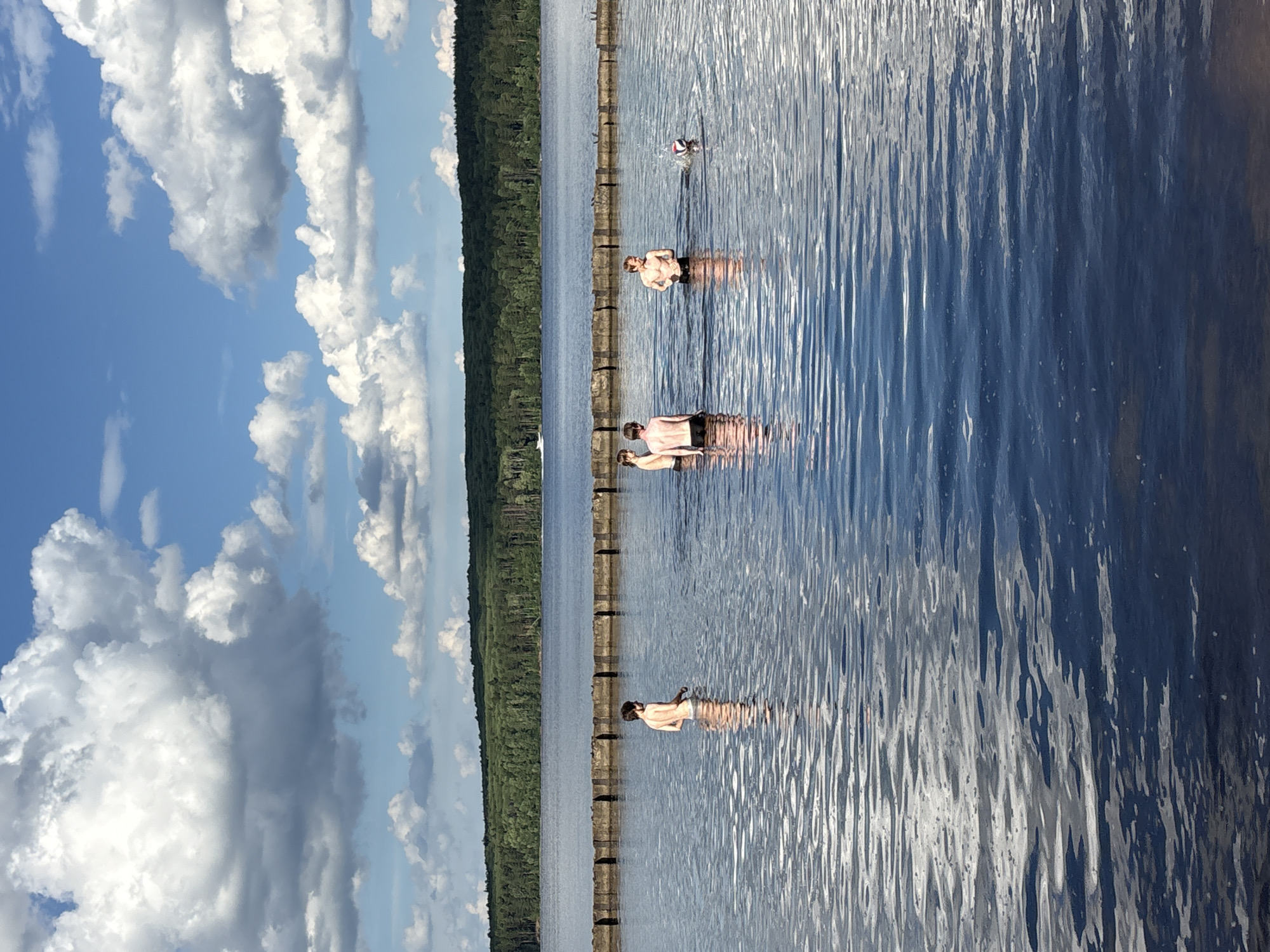 In a serene lake setting, four individuals are immersed in the water, enjoying a leisurely day. The scene is bathed in the bright light of midday, with fluffy white clouds dotting a clear blue sky, indicating fair weather. The lake, with its calm, rippling surface, reflects the surrounding verdant forest that lines the distant shore. A submerged wooden structure, likely a pier or a barrier, runs across the lake, serving as a visual marker and a point of reference for the people in the water.
Two individuals are closer to the foreground, wading in the water, their torsos visible above the surface. They appear relaxed, engaged in conversation or simply enjoying the cool water. Further in the distance, two more individuals are visible. One is partially submerged, resting in the water, while another is further out, possibly playing with a ball, as suggested by a round object near them. The overall atmosphere is one of tranquility and outdoor recreation. The orientation of the image is landscape, with the lake and the individuals filling the frame.