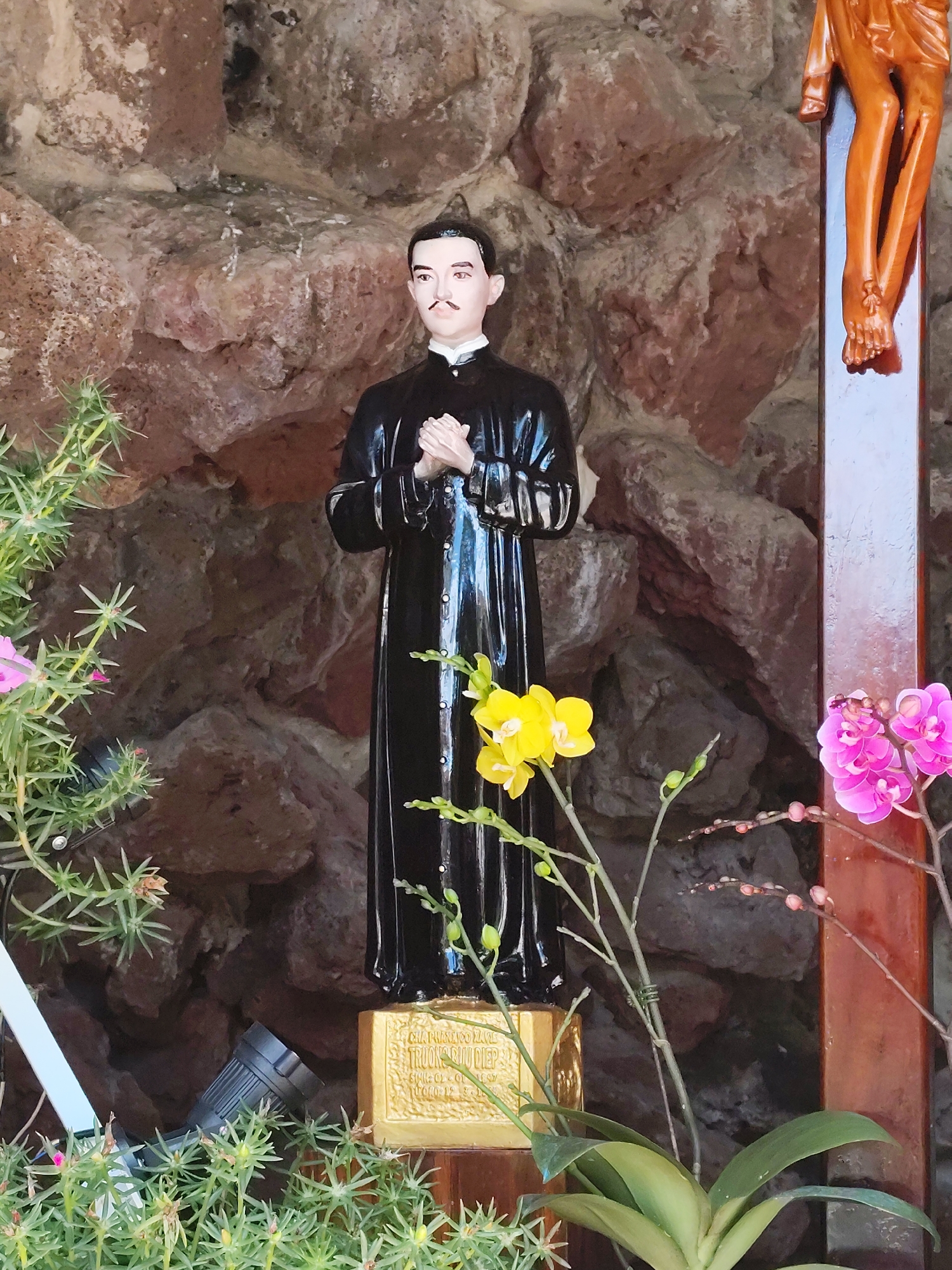 In Da Lat, Vietnam, a statue of a priest stands in front of a rough-hewn stone wall. The priest, depicted in a black robe with his hands clasped, is positioned on a gold pedestal. The scene is adorned with flowering plants, including orchids in vibrant yellow and pink hues, and delicate green foliage. To the right of the statue, part of a wooden crucifix, showing the legs of Christ, is visible. The environment suggests an outdoor shrine or a religious garden setting.