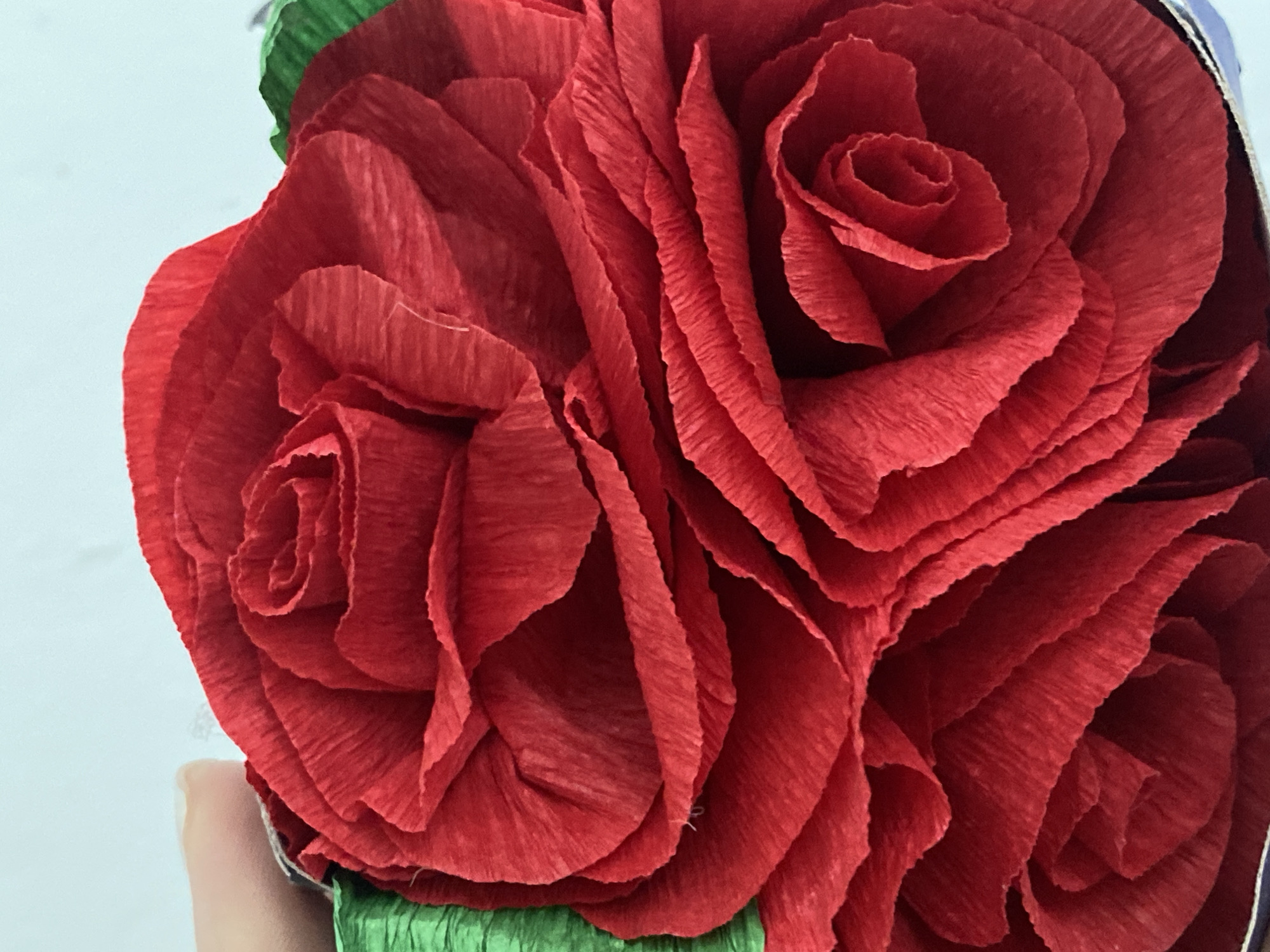 A close-up shot shows a bouquet of three handmade red crepe paper roses with green crepe paper stems. The roses are arranged tightly together, filling the frame. The texture of the crepe paper is clearly visible, with distinct lines and folds creating a realistic appearance. A light-colored surface is visible in the background to the left. There is no information about the city of Sochi, Russia, in the image.