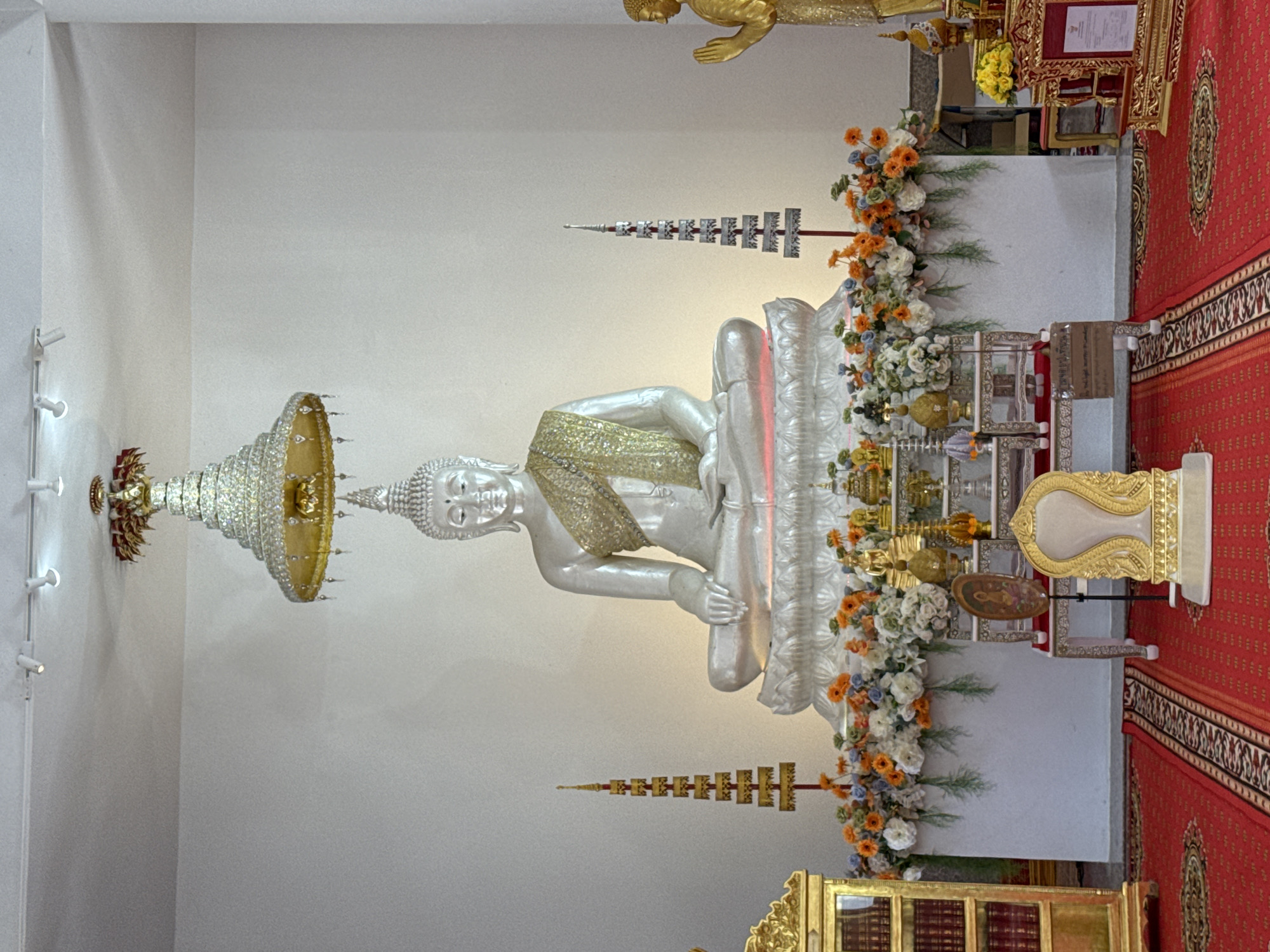 The image depicts the interior of a Buddhist temple in Thep Krasatti, Thailand, featuring a large, ornate white Buddha statue seated on a raised platform. The Buddha is adorned with a golden sash. Above the statue, a tiered, crystal-like umbrella-shaped structure hangs from the ceiling. The platform is decorated with an abundant display of white, orange, and blue flowers, along with various golden religious artifacts. To the right of the Buddha, a smaller golden statue of a person with hands clasped in a prayerful gesture stands. In front of the platform, a red carpet with intricate patterns covers the floor. A golden, ornate chair with a white cushion is positioned on the left side of the foreground, and a tall, narrow golden shelf unit filled with books is located to the far left. The walls of the temple are white, and overhead track lighting illuminates the scene.