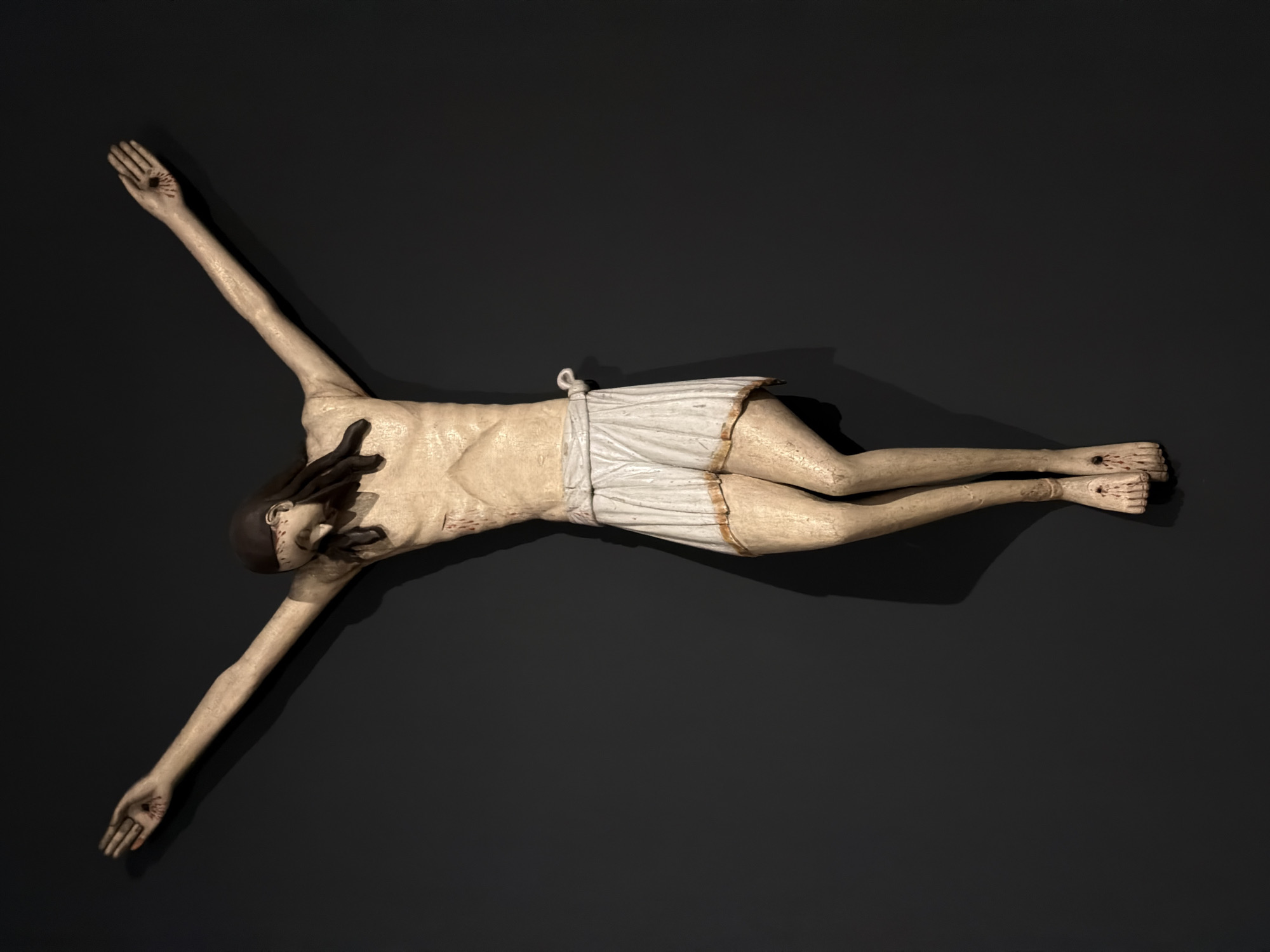 A high-angle shot depicts a light-skinned sculpture of a crucified figure, lying diagonally against a solid dark gray background. The figure has its arms extended outward in a crucifix position. The hands have what appears to be nail holes in the center of the palms. The lower body is covered by a white cloth tied at the waist. The feet are crossed, one over the other, with visible nail wounds. The sculpture appears to be depicting Jesus Christ. The sculpture is from the Perm Art Gallery in Perm, Russia.
