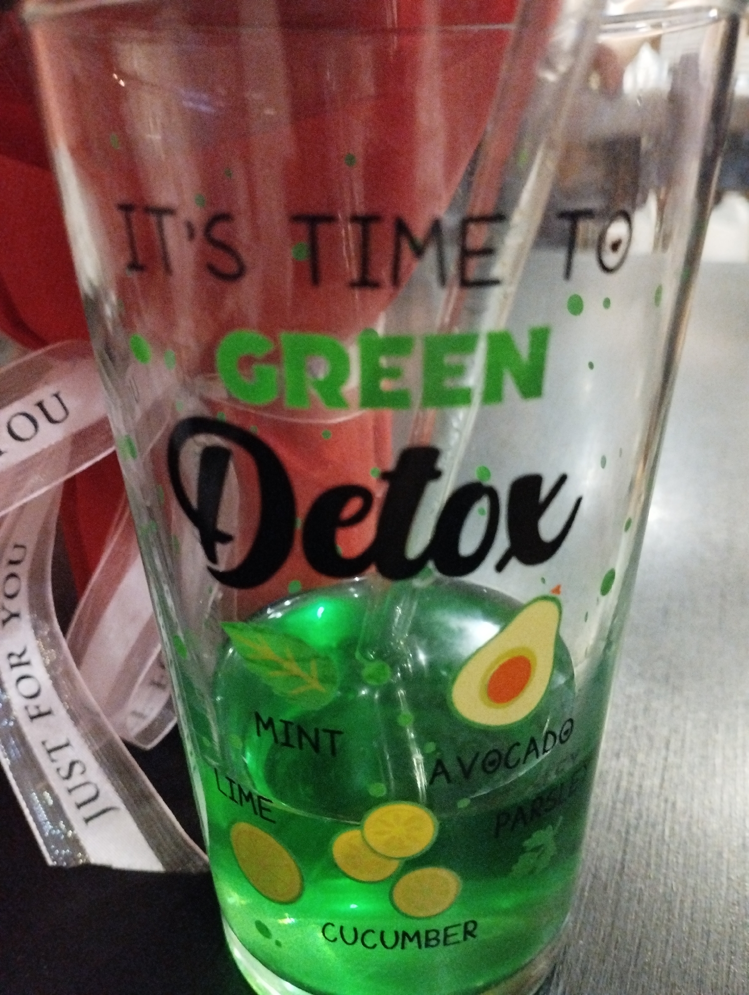 A close-up shot shows a clear glass that is decorated with the words "IT'S TIME TO GREEN Detox" printed on its exterior. The word "GREEN" is colored green, while "Detox" is in black. The glass contains a green liquid, possibly a detox drink, which is visible through the clear glass. The glass is also adorned with cartoonish images of ingredients such as mint, lime, avocado, parsley, and cucumber, with their respective names printed next to them. A clear straw is partially visible inside the glass. In the background, there's a red object and a white ribbon printed with the words "JUST FOR YOU". The surface beneath the glass appears to be a table with a gray or silver color. The photo appears to have been taken indoors. The location in the city of Burgas, Bulgaria cannot be determined from the provided image.