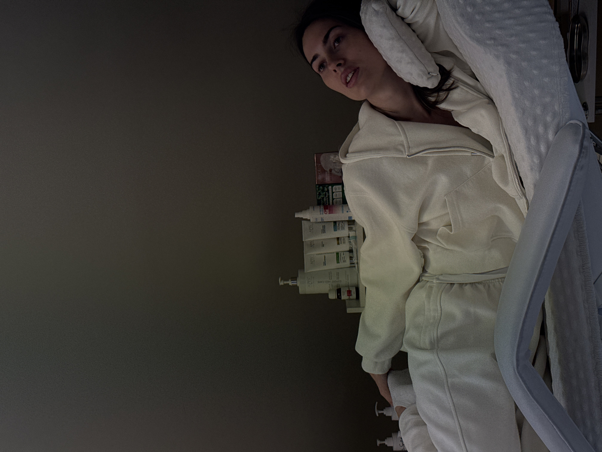 A woman lies on a white, cushioned chair. She is wearing a light-colored sweatsuit with a zippered hoodie. A shelf with various bottles and containers is visible near her head. The background is a dark, solid color, suggesting a dimly lit room. The scene is in the city of Unknown, Unknown.