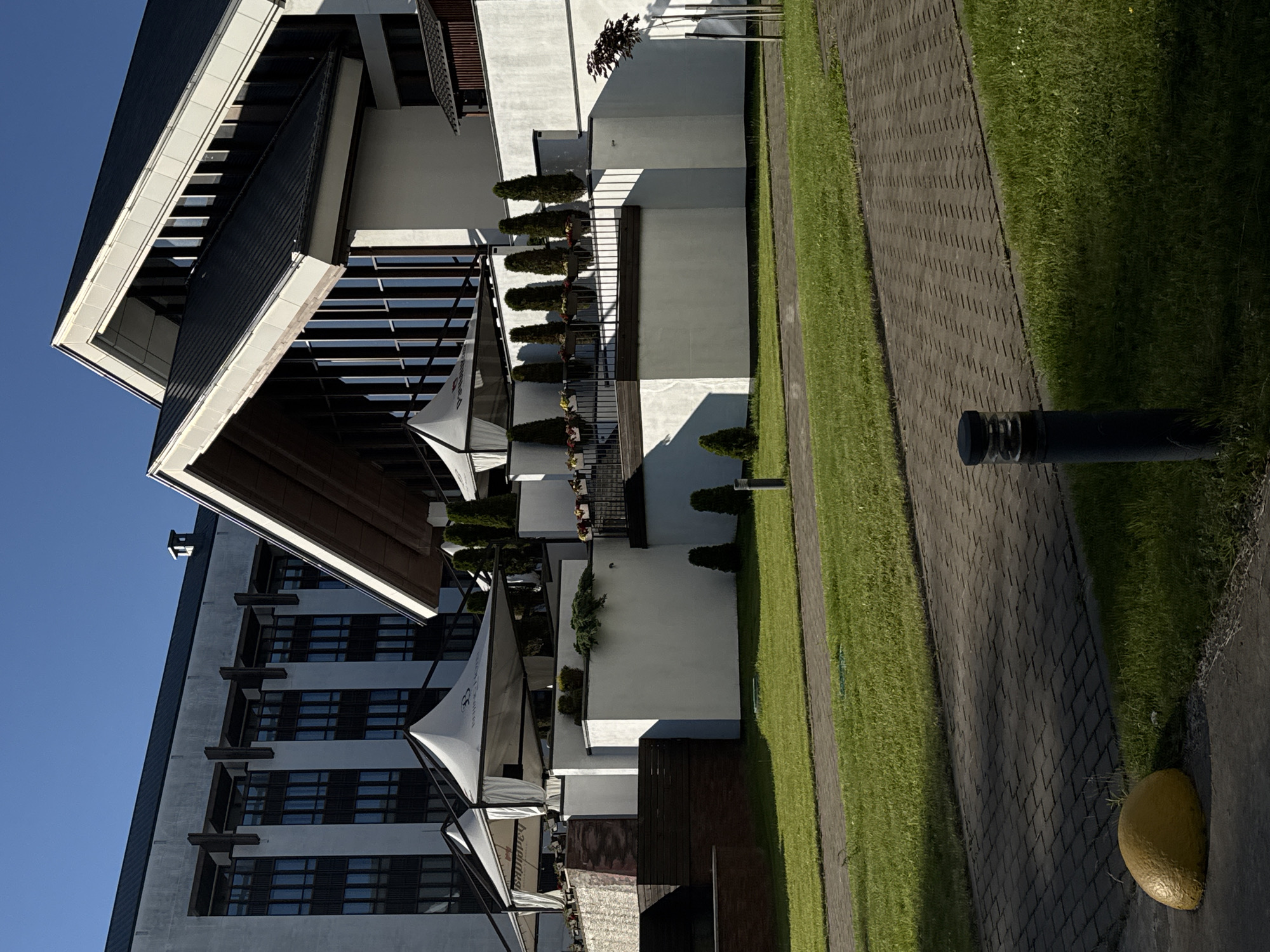 The image shows a modern building with a white facade and a dark, sloped roof. In the foreground, there is a neatly mowed green lawn and a paved walkway. A yellow bollard is visible on the left side of the walkway, and a dark, cylindrical lamp post is on the right. The building features large windows, some of which are reflecting the blue sky. White canopies are set up on a terrace area, and there are potted plants and decorative cypress trees along the building's perimeter. The overall scene suggests an outdoor seating or relaxation area of a commercial establishment, possibly a hotel or restaurant. The architectural style is contemporary. The provided crop images are consistent with this description, focusing on different parts of the building and its surroundings. The building appears to be located in an urban or suburban environment.