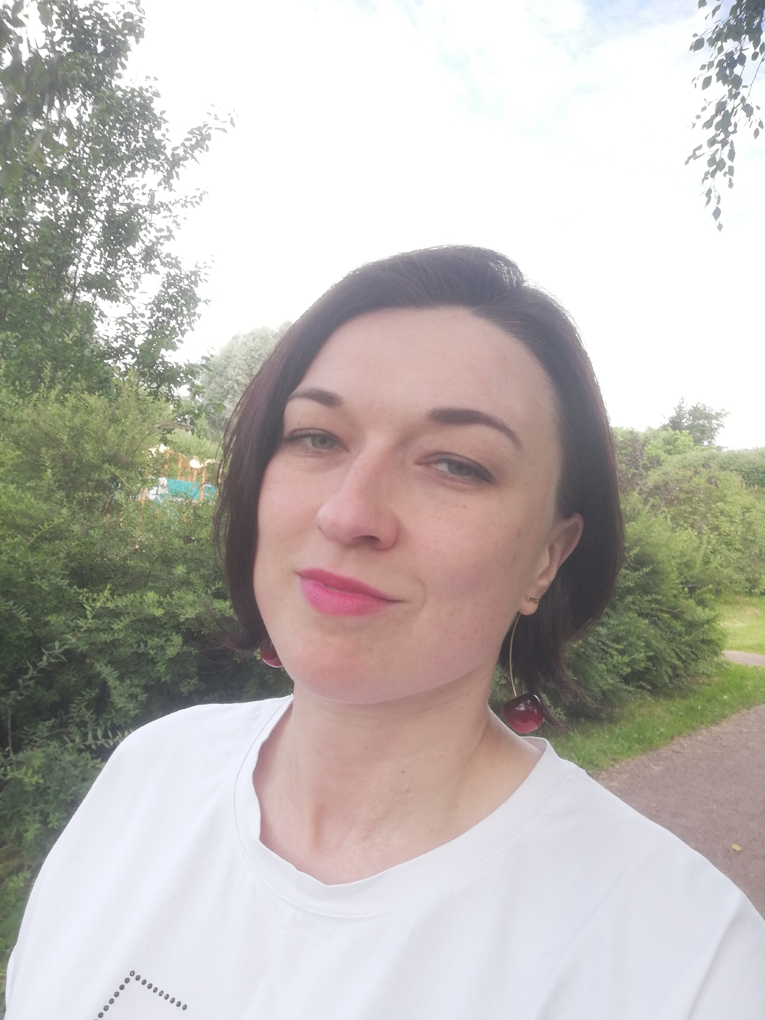 The image is a selfie taken outdoors with a woman in the foreground. She has dark hair, fair skin, and is wearing a white t-shirt. She has bright pink lipstick on and is wearing large cherry-shaped earrings. Behind her, there is lush green foliage and a pathway, suggesting a park or natural setting. The sky is overcast and bright, indicating a daytime shot. The overall mood appears casual and relaxed. There is no visible text in the image. The context indicates the location is Kolpino, Russia.