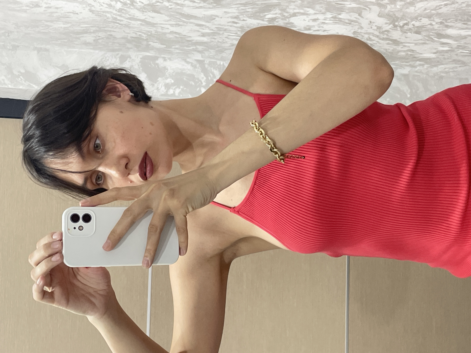 A woman with dark hair and brown eyes takes a selfie using a white smartphone. She is wearing a red ribbed top with thin straps and a gold bracelet on her left wrist. The background consists of a textured white surface at the top and a plain beige surface at the bottom. The location is not specified, and the image does not provide sufficient information to determine if it was taken in Izhevsk, Russia.
