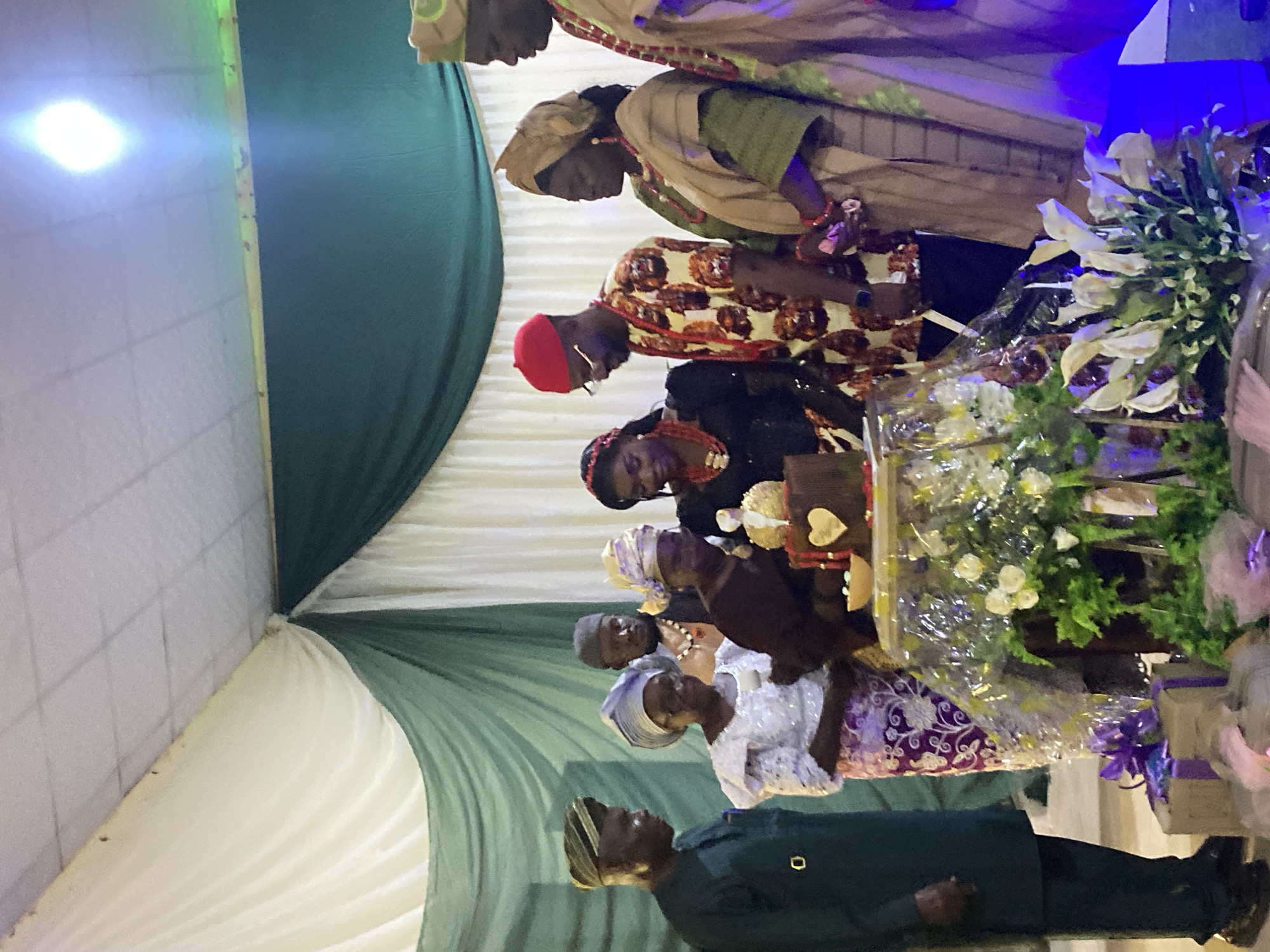 A group of people are gathered around a table. On the table, there are flowers and gifts. The people are dressed in traditional clothing. The environment appears to be a venue decorated with green and white fabrics. The location is Vom, Nigeria.