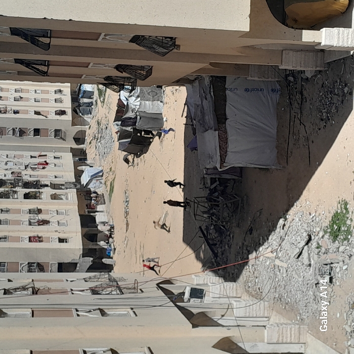 The image captures an outdoor scene in Khan Yunis, Palestinian Territory, centered on a large, unpaved dirt and sand area surrounded by multi-story residential buildings. These concrete structures exhibit numerous windows, many with external metal grates or shutters, some of which appear damaged or askew. Clothes are visible hanging from windows on multiple floors.
The open ground is heavily strewn with concrete rubble, twisted metal, and various debris. Several makeshift shelters and tents, constructed from tarpaulins and fabric, are set up across this area; one prominent shelter on the right displays a visible UNICEF logo.
In the mid-ground, five individuals are in motion. A figure in a light-colored shirt is depicted in a throwing posture, holding a light, flat object. Nearby, another individual in a dark shirt runs. A third figure in a blue shirt is seen running towards the right side of the frame. Two additional figures, one in a red shirt and one in light clothing, are positioned further left. In the background, near a distant building and another tent structure, a small group of approximately three to five individuals is gathered. Long shadows are cast across the ground from the surrounding structures and foreground objects. Overhead wires or cables extend across the left side of the foreground. A "Galaxy A14" watermark is present in the bottom left corner.