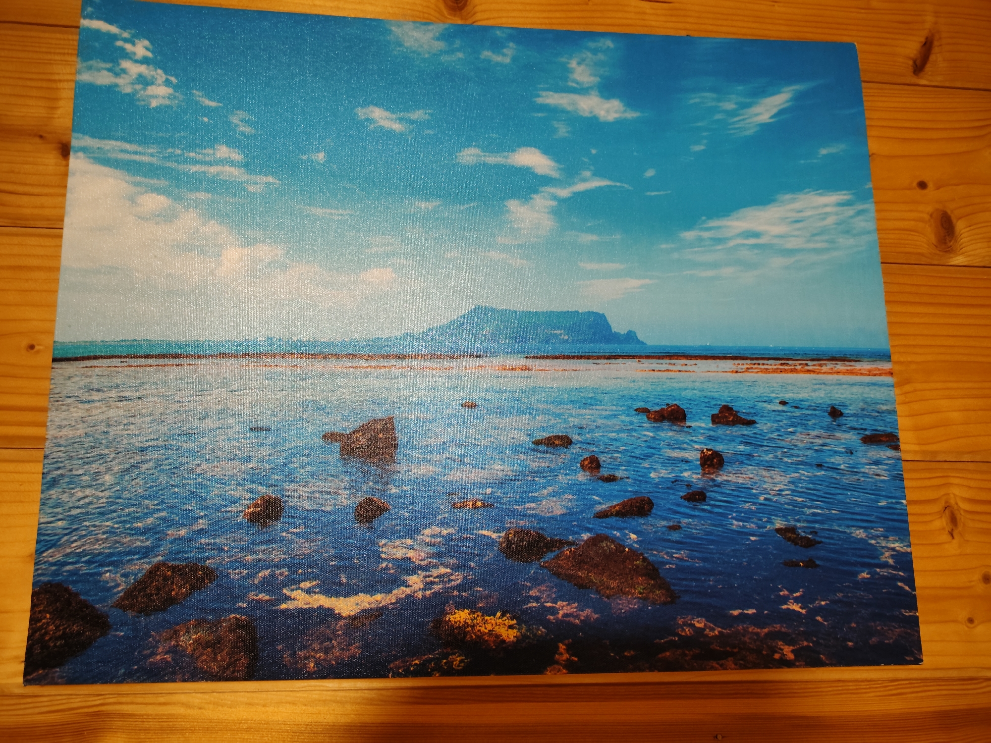 The image displays a picturesque seascape. In the foreground, dark, wet rocks are scattered in shallow, clear blue water, some partially submerged. The water's surface reflects the sky and is disturbed by gentle ripples, revealing patches of orange and yellow seaweed or algae below. The middle ground features a wider expanse of this blue water, with more rocks and the shoreline visible in the distance. Beyond the water, a low, verdant, and somewhat mountainous island or peninsula stretches across the horizon. The sky above is a vibrant blue, dotted with fluffy white clouds. The overall scene is serene and natural, depicting a coastal landscape. There are no people or man-made structures visible in the image, suggesting a remote or undeveloped location. The image is a photograph of a print, likely a canvas or poster, placed on a wooden surface composed of planks with visible grain and knots, typical of a table or wall. The lighting suggests it is daytime, with the sun illuminating the scene and creating reflections on the water. The location is described as "Фестивальное Снт, Russia."