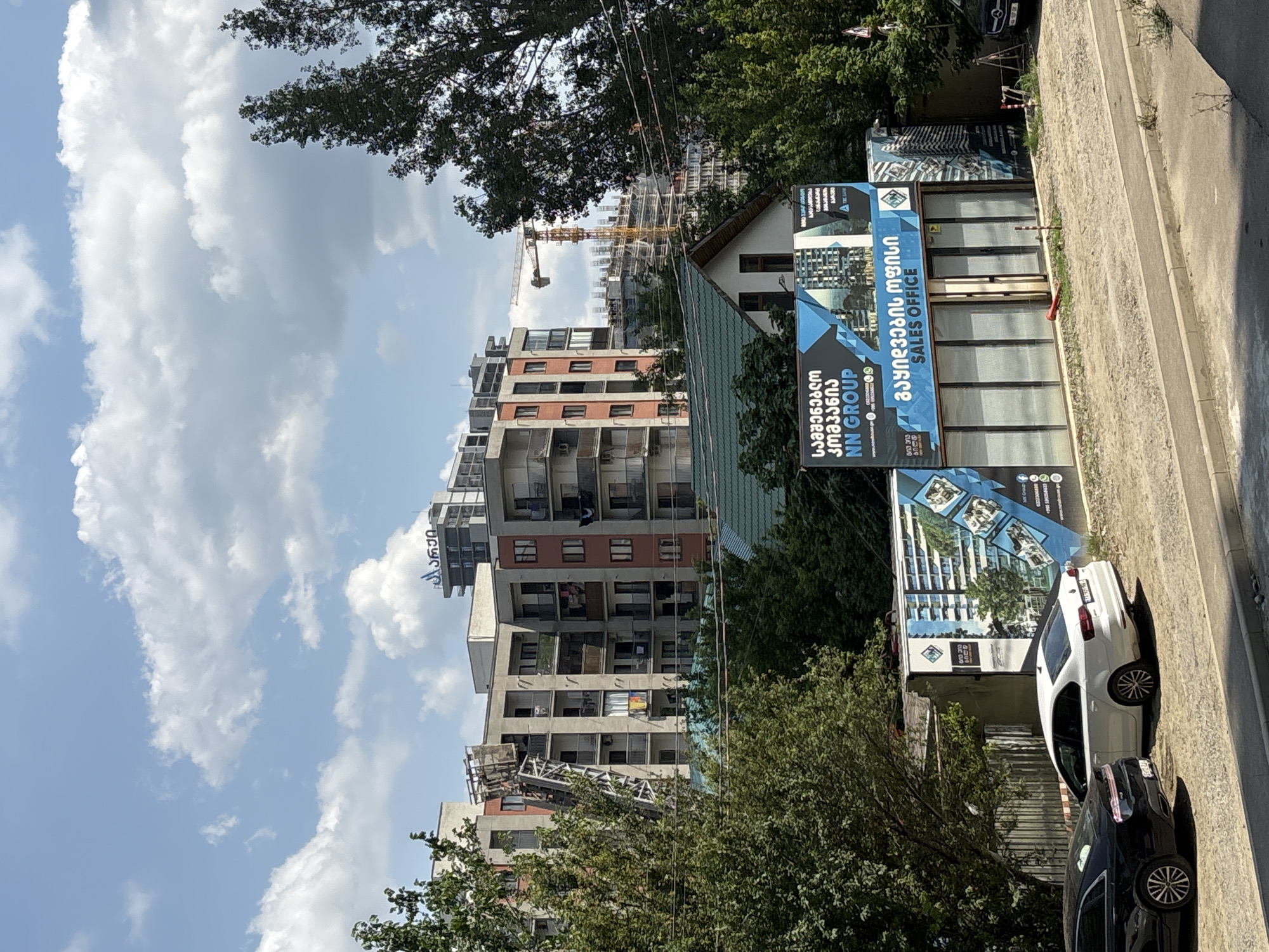 The image depicts a street scene in Tbilisi, Georgia, under a partly cloudy sky. In the background, several multi-story apartment buildings are visible. One building on the left has an external elevator or construction lift attached to its facade. Further in the background, a yellow construction crane rises above other buildings under construction. In the foreground, a sales office for "NN GROUP" is situated behind a row of trees. The sales office has a large glass storefront and is adorned with a prominent advertisement featuring images of modern apartment complexes. Two cars are parked in front of the sales office, one black and one white. The area appears to be a developing urban or suburban environment.