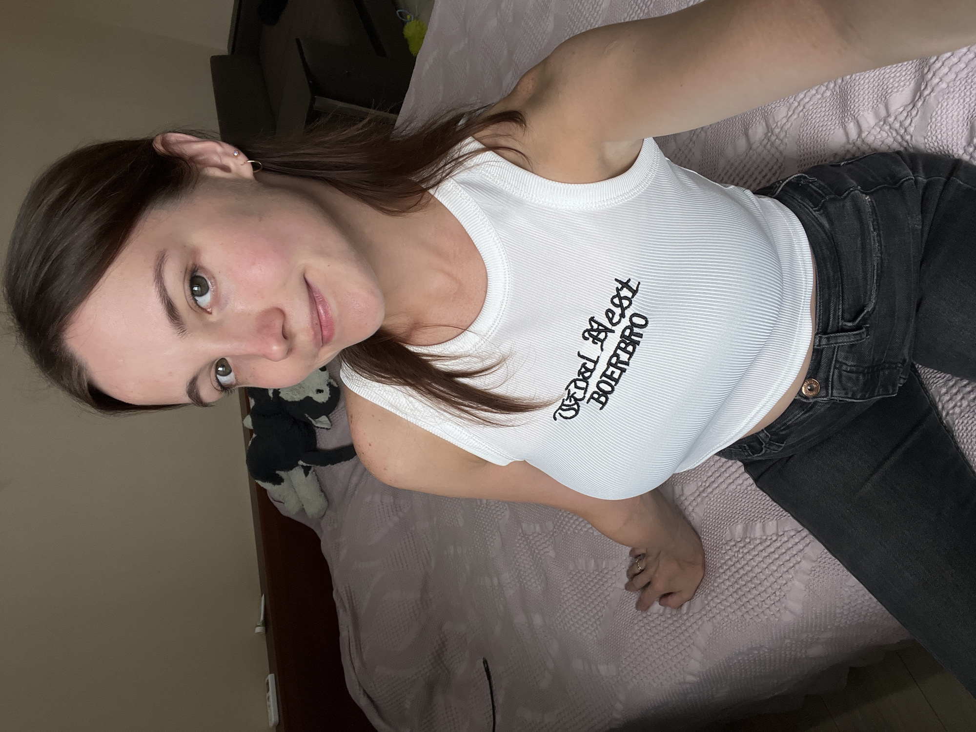 A young woman with brown hair and green eyes is sitting on a bed, facing the camera. She is wearing a white ribbed tank top with black text that reads "Wood Nest BOERBRO" and dark wash denim jeans. The bed is covered with a light pink textured comforter. In the background, there is a wooden headboard and two stuffed animals, one black and white and the other grey and white. The room appears to be a bedroom. There are no visible landmarks that would indicate the location is in the city of Rozhdestveno, Russia.