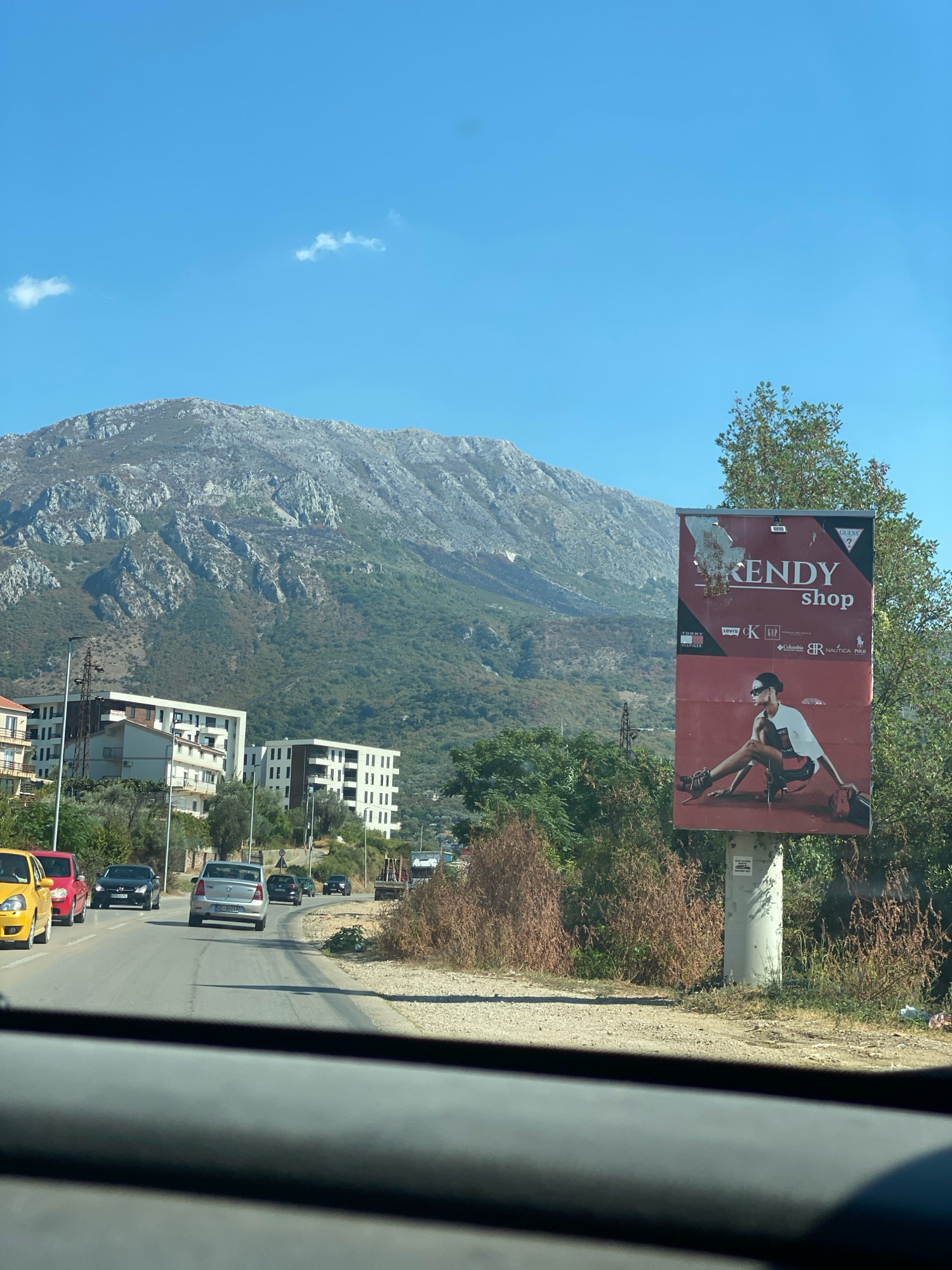This is a ground-level, exterior, eye-level shot looking forward from a vehicle through the windshield. The scene is a paved road with cars driving on it, with a billboard on the right side and buildings and a mountain in the background.
The road is asphalt with white lane markings. Several cars are visible on the road, including a yellow car on the left, a red car behind it, a black sedan, a silver sedan with a license plate, and other vehicles further down the road. The billboard is red and displays an advertisement for "KENDY shop" featuring a model. The billboard is mounted on a concrete pole.
In the background, there are several buildings, including modern multi-story apartment buildings with white exteriors and dark windows. Behind the buildings, a large, rocky mountain with sparse vegetation dominates the landscape under a clear blue sky with a few scattered clouds. Trees and dry brush are present on the right side of the road, near the billboard.
The location is identified as Čeluga, Montenegro. The image appears to have been taken during the daytime, likely in the morning or afternoon, given the clear sky and lighting. There are no people clearly visible in the image.