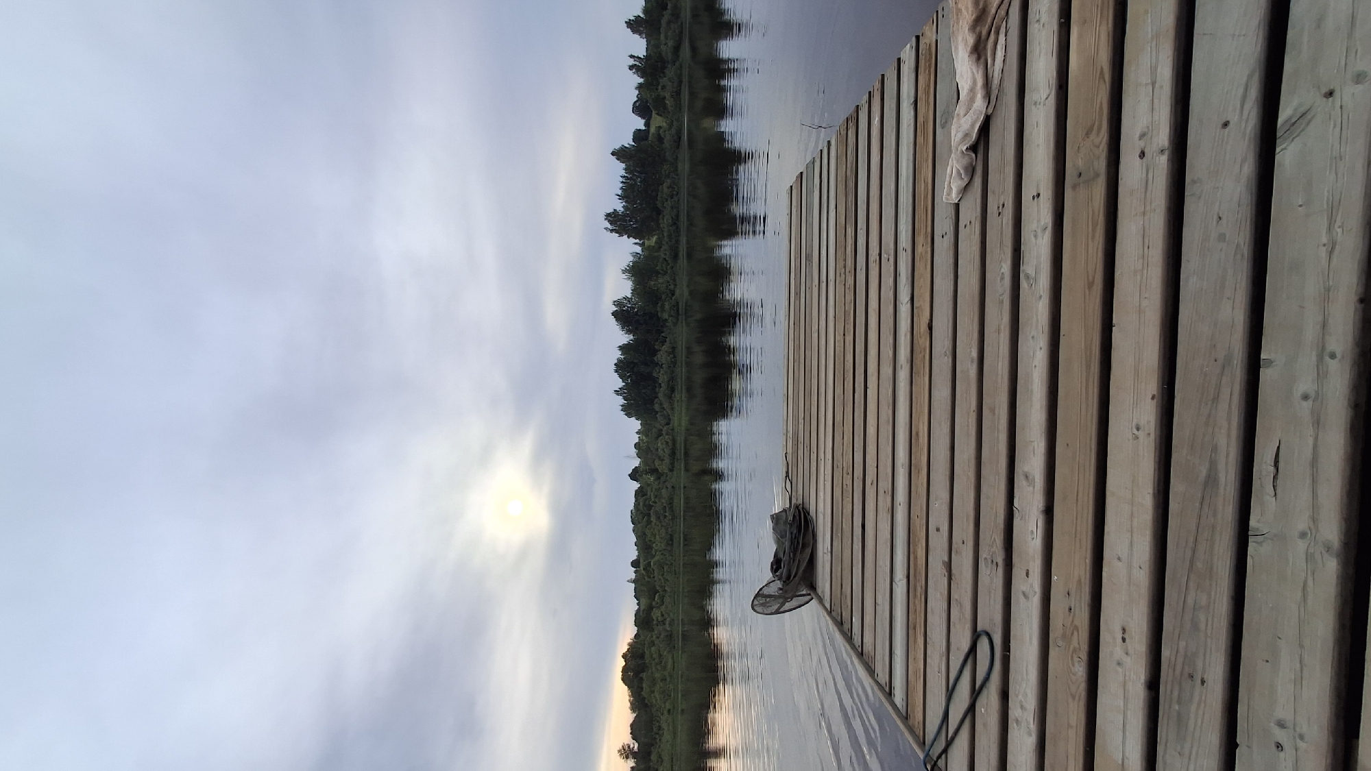 The image depicts a wooden pier extending into a calm body of water, reflecting the overcast sky and a distant, tree-lined shore. The sun is partially visible through the clouds, casting a soft glow. On the pier, a fishing net and a folded cloth are present. The scene appears to be a natural landscape, possibly a riverbank or lakefront, located in the vicinity of Sitno, Russia. There are no people or animals visible in the image.