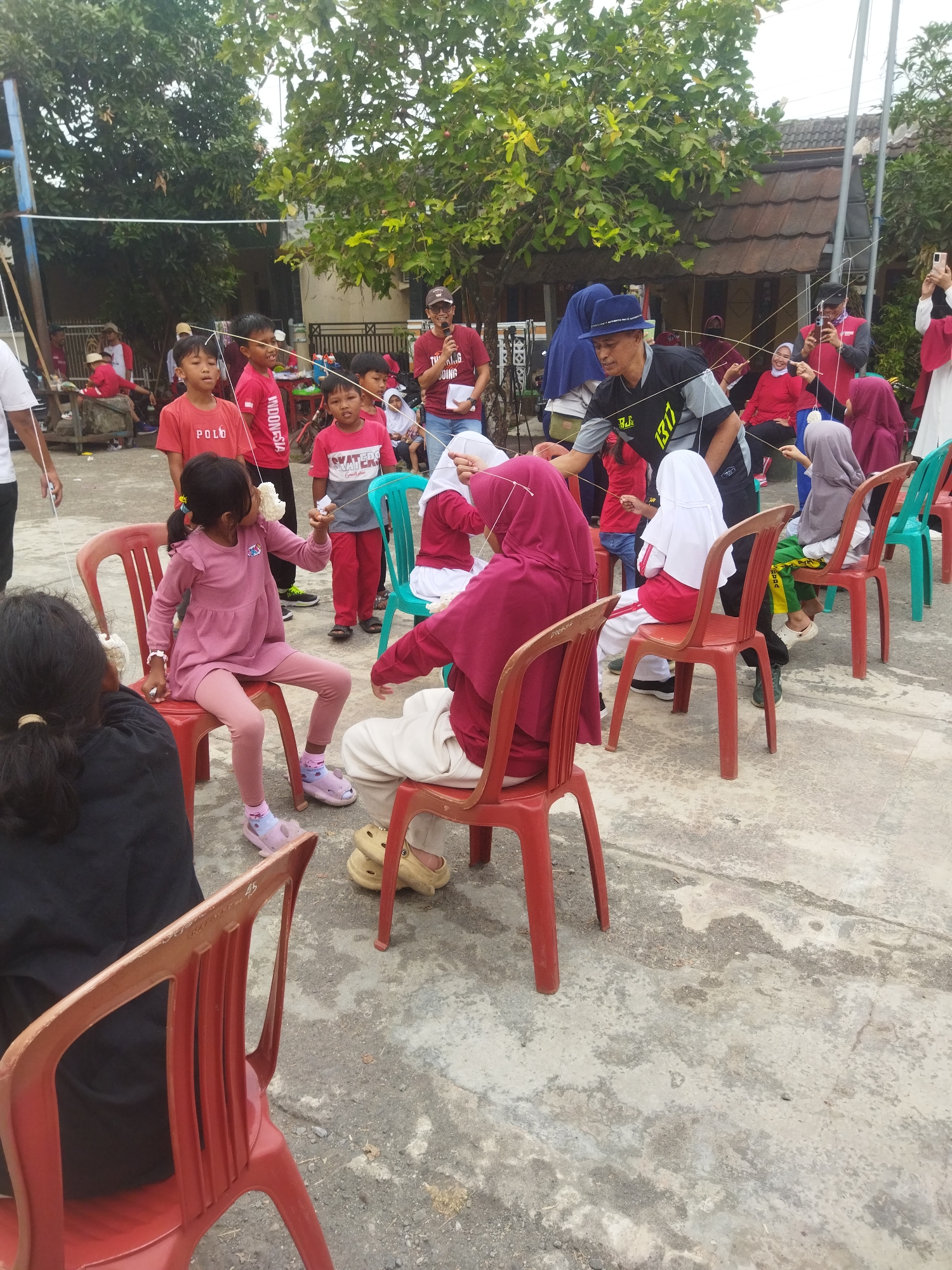In Banjar, Indonesia, children and adults participate in an outdoor game involving eating crackers suspended by strings. Several children are seated on red and green plastic chairs, each attempting to eat a cracker dangling from a string held by adults or older children. One girl, seated on a red chair, is actively eating her cracker, holding the string with her hand. Another child, also on a red chair, is visible in the foreground. Adults stand behind the seated children, managing the strings and observing the game. The environment appears to be a concrete courtyard with trees and buildings in the background. The event seems to be a community gathering or celebration.