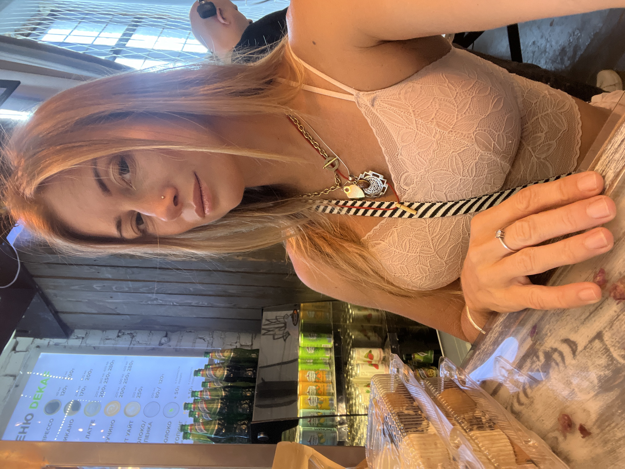 A young woman with blonde hair, wearing a light-colored lace bralette and several necklaces, poses for a selfie at a counter. To her left, there are stacks of pastries in plastic containers. Behind her, a refrigerated display case is stocked with bottled drinks. A menu board is visible on the left wall, displaying prices in Russian rubles. The setting appears to be a cafe or bar in Moscow, Russia, given the Cyrillic script on the menu and the overall ambiance.