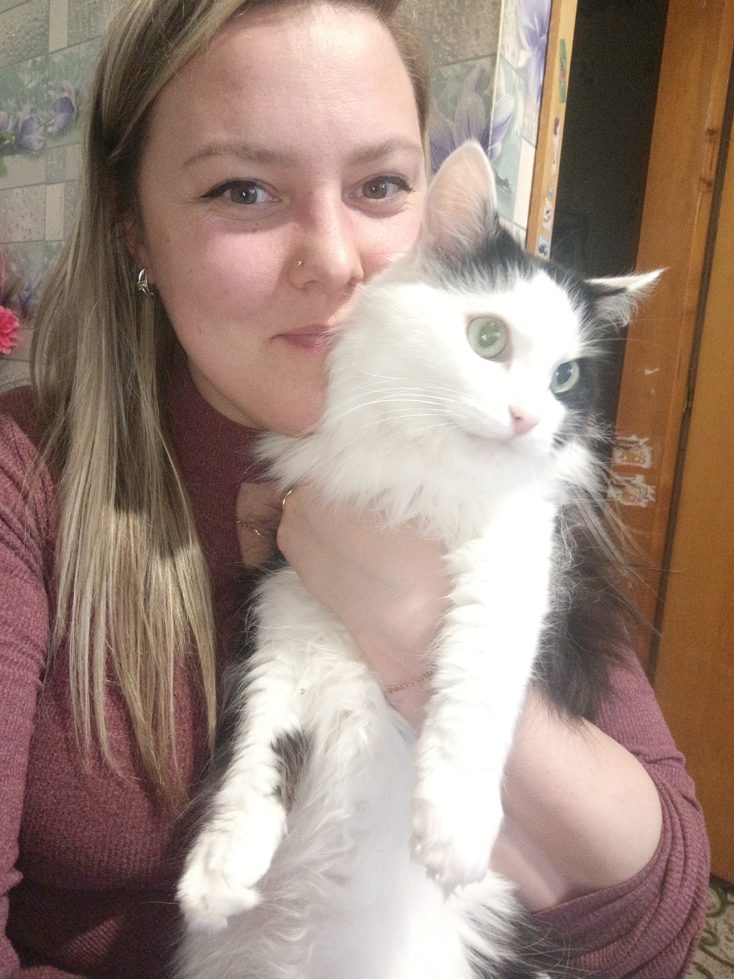 A woman is holding a black and white cat indoors in what appears to be a home setting in Марковский, Russia. The image is a close-up, likely a selfie, capturing both subjects in a warm, affectionate pose.
The woman has light skin, long, straight blonde or light brown hair, and is wearing a maroon or burgundy ribbed long-sleeved top. She has a gentle smile, brown eyes, and is looking directly at the camera. Notable details include a small gold stud nose piercing on her left nostril and a silver hoop earring with a green embellishment in her left ear. A thin gold chain bracelet is visible on her right wrist.
She is cradling a fluffy, long-haired cat, predominantly white with distinct black patches on its head (around the ears and one eye) and body. The cat possesses striking green eyes, one of which appears to have a stronger reflection or a slightly different hue. Its long white whiskers are prominent. The cat is looking slightly to the right, away from the camera, with a neutral to curious expression.
The background features a wall adorned with floral wallpaper, depicting purple or lilac flowers on a lighter base. To the far right, a wooden door frame or door is visible. On this wooden surface, there is a blurry label or sticker with text that is unreadable but appears to be in an Asian script. The overall lighting is soft, typical of an indoor environment. The woman's posture and smile suggest a loving bond with her pet.