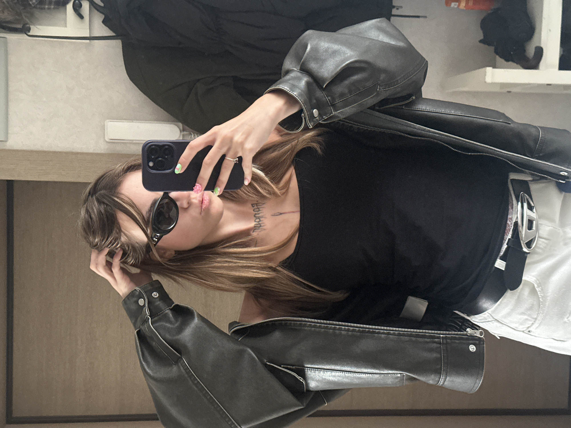 In Moscow, Russia, a young woman is taking a selfie in a hallway. She is wearing a black tank top, a black leather jacket, and white pants. She has on black sunglasses and is holding a smartphone in her right hand, which she is using to take the photo. Her left hand is touching her hair. She has tattoos on her chest and a ring on her right ring finger. Her nails are painted with green and pink designs. A black jacket is hanging on a rack behind her, and a shelf with various items is visible to the right. The walls appear to be a light color, and a wooden door is on the left side of the frame.