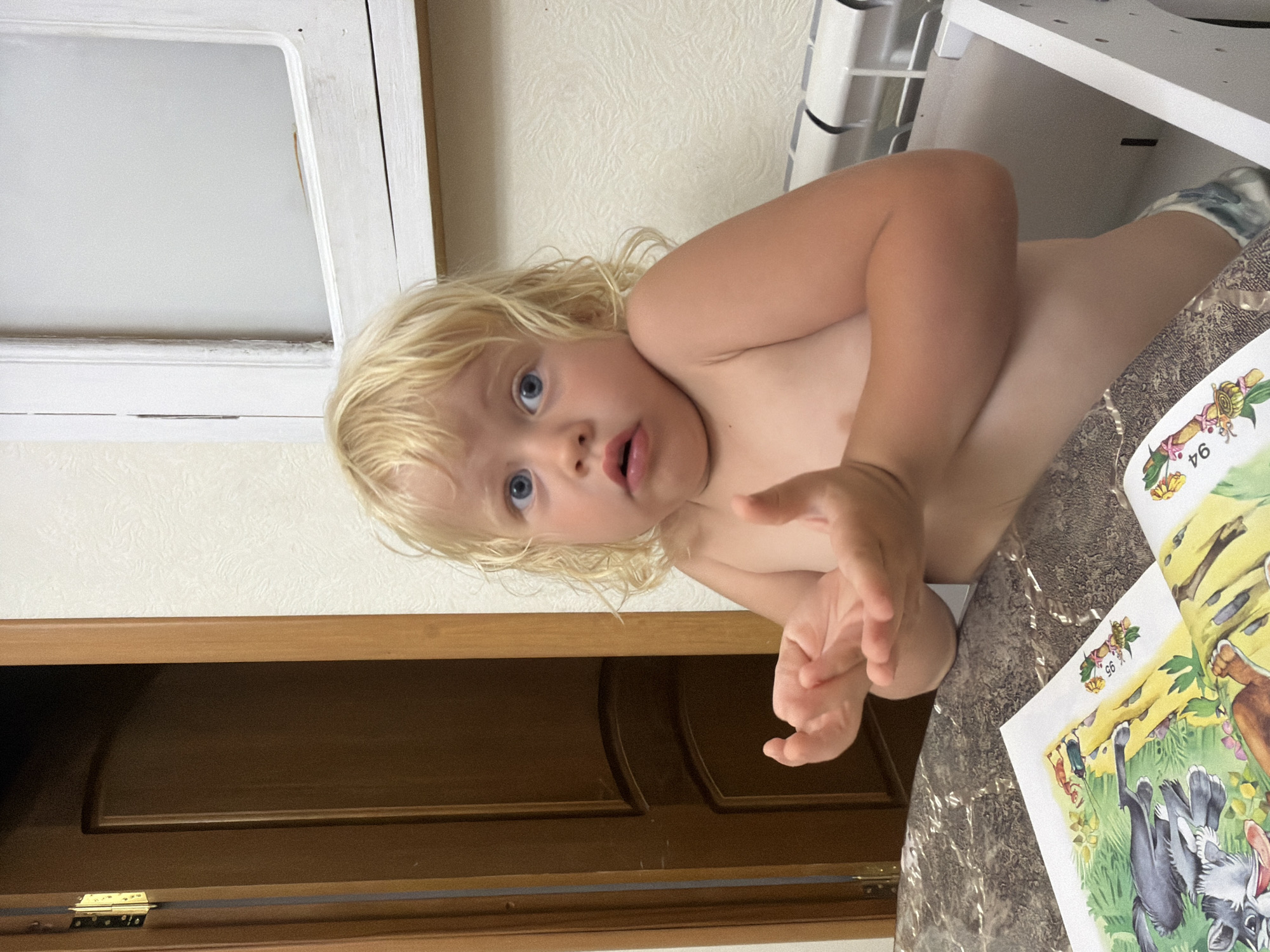 A close-up, rotated shot shows a young child with blonde, curly hair and blue eyes lying on their stomach, looking up at the camera. The child appears to be indoors, in what seems to be a home environment. The background includes a textured wall, a white radiator, and a wooden cabinet. The child is wearing socks, one of which has a patterned design.
To the right of the child, a colorful children's book is open. The illustrations depict animals, including a rabbit and a squirrel, in a grassy setting. There is a small tag on the page with the number "96" printed on it.
The child's expression is one of surprise or curiosity, with their mouth slightly open and eyes wide. Their hands are raised, as if reaching out or gesturing. The lighting suggests it could be daytime, but no specific time is indicated. The location is noted as Sochi, Russia. The scene is intimate and suggests a moment of play or interaction between the child and whoever is taking the photograph.