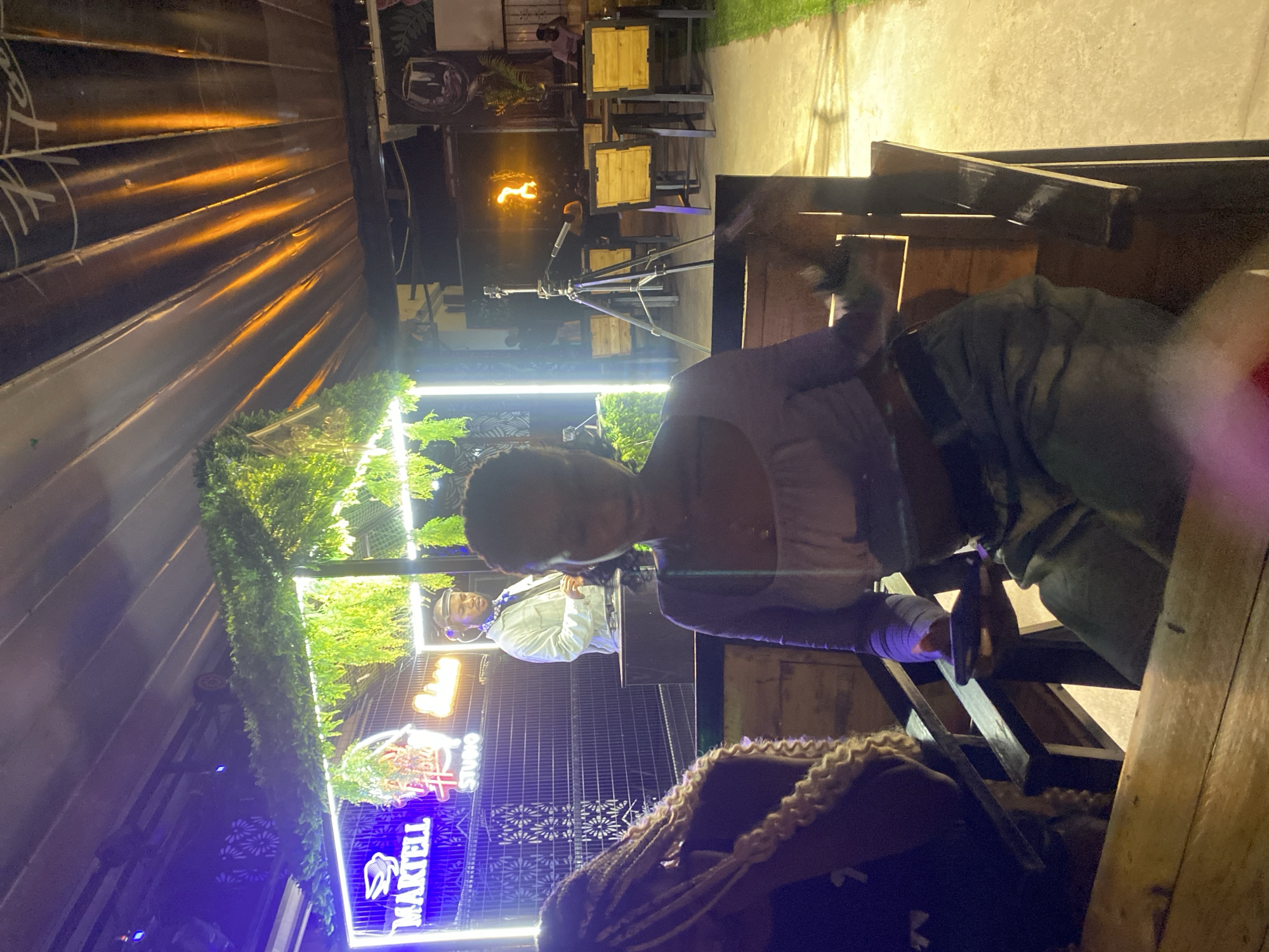 The image depicts an outdoor evening scene at a venue in Howayi, Nigeria. In the foreground, a woman is seated at a wooden table, looking down at her phone. To her left, another woman with blonde braids is partially visible. In the background, a DJ wearing headphones and a light-colored jacket stands behind a DJ booth, illuminated by neon signs that spell out "MARTELL" and "HOWAYI STUDIO". The venue appears to be an open-air space with wooden chairs and tables, a grassy area, and decorative greenery. A tripod with a camera and microphone is set up to the right, facing a wall where a fire performance is occurring, with flames illuminating the area. The ceiling is covered with corrugated metal, reflecting warm orange light.