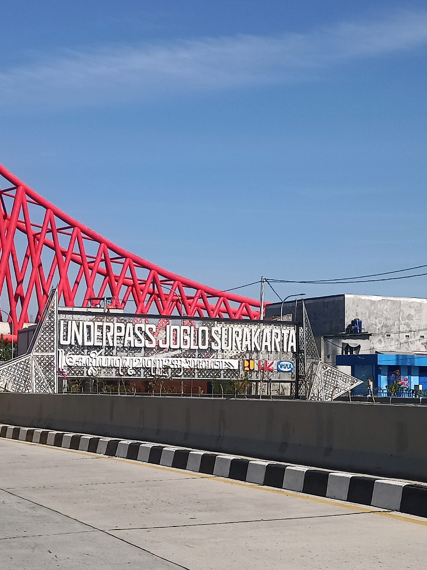 The image captures an outdoor scene in Surakarta, Indonesia, featuring a prominent underpass entrance. A bright red, intricate metal structure, likely part of the underpass's architecture, spans the upper left portion of the frame. Below this, a large sign reads "UNDERPASS JOGLO SURAKARTA" in white, bold lettering. Beneath this main text, a line of text in what appears to be Javanese script is visible. The sign is framed by decorative white panels with an intricate geometric pattern. To the right of the underpass entrance, a concrete building with a somewhat rough finish is visible. Further to the right, a brightly colored blue structure with some people and plants can be seen. The foreground shows a road with a concrete barrier, marked with alternating black and white blocks and a yellow line. The sky is clear and blue, suggesting a sunny day.