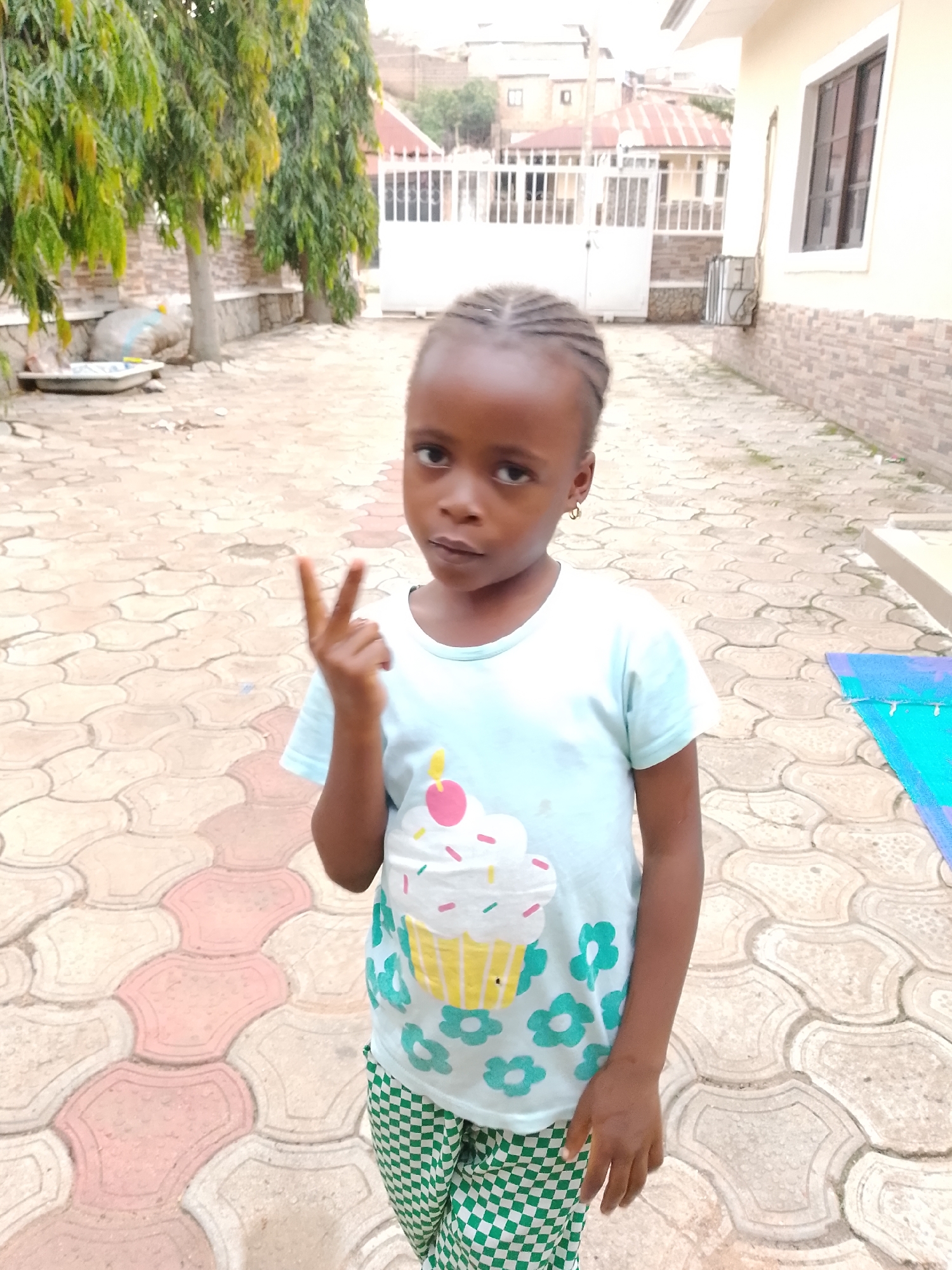 A young girl is standing outside in a paved area. She is wearing a light blue t-shirt with a cupcake design and green and white checkered pants. She is holding up two fingers in a peace sign. In the background, there is a building with a white gate, trees, and houses. The ground is paved with interlocking concrete tiles in shades of beige and pink. There is also a blue mat on the ground to the right of the girl.