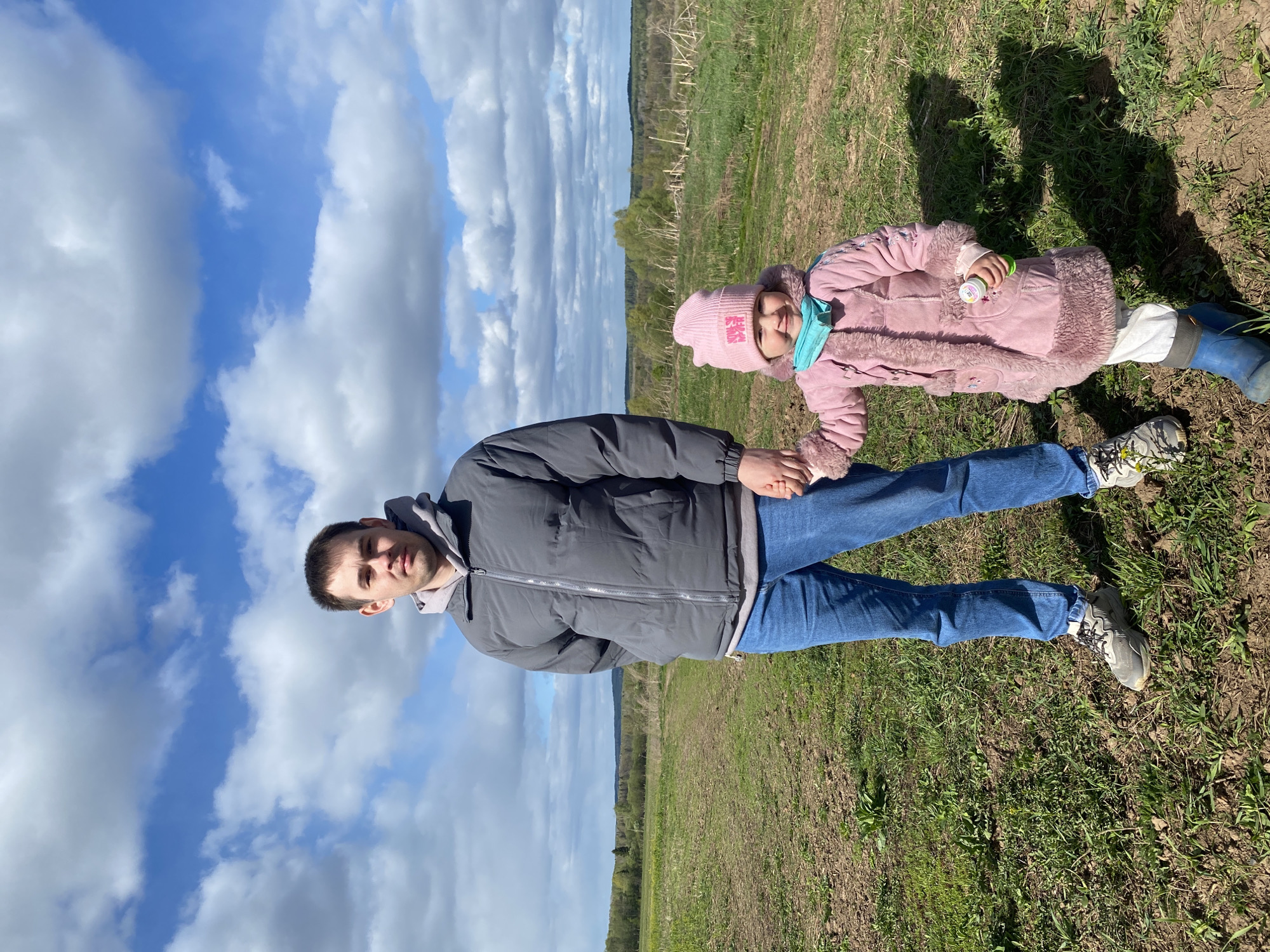 A man and a young child are lying in a grassy field under a partly cloudy sky. The man is wearing a gray puffer jacket, jeans, and sneakers. The child, lying beside him, is wearing a pink hat, a pink coat, and blue rain boots. The child is holding a small container. The background includes a distant treeline. The location is likely rural, possibly near the city of Дедушкино, Russia.
