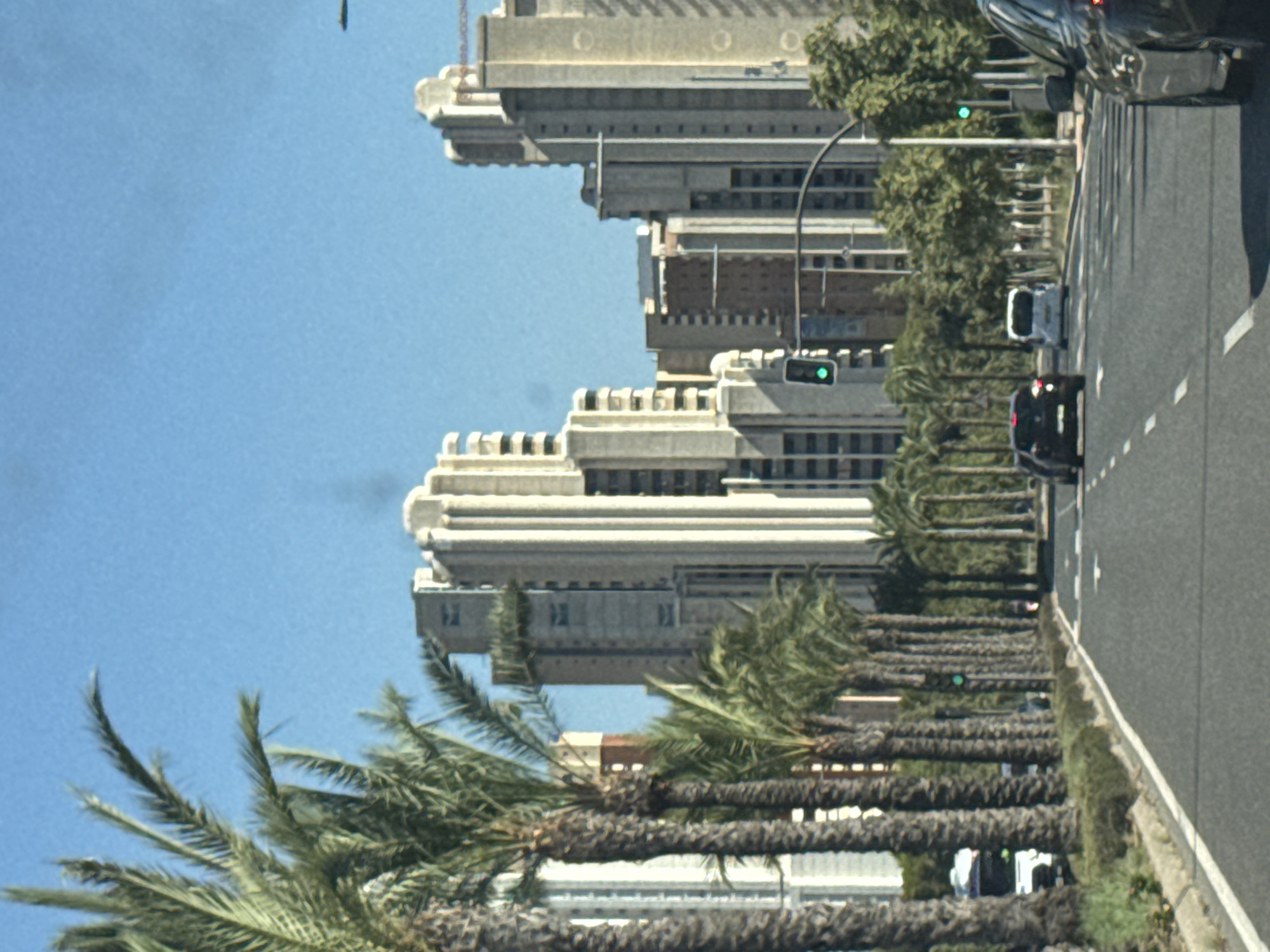The image depicts a street scene in Benidorm, Spain, characterized by a wide asphalt road lined with palm trees. Tall, modern buildings, likely hotels or apartment complexes, dominate the skyline in the background. Several cars are visible on the road, including a dark-colored SUV in the foreground to the right, and a black sedan and a white van further ahead. Traffic lights are present, with a green light illuminated on one signal. The sky is clear and blue. The environment suggests a sunny day in a developed urban area with a tropical or Mediterranean feel due to the palm trees.