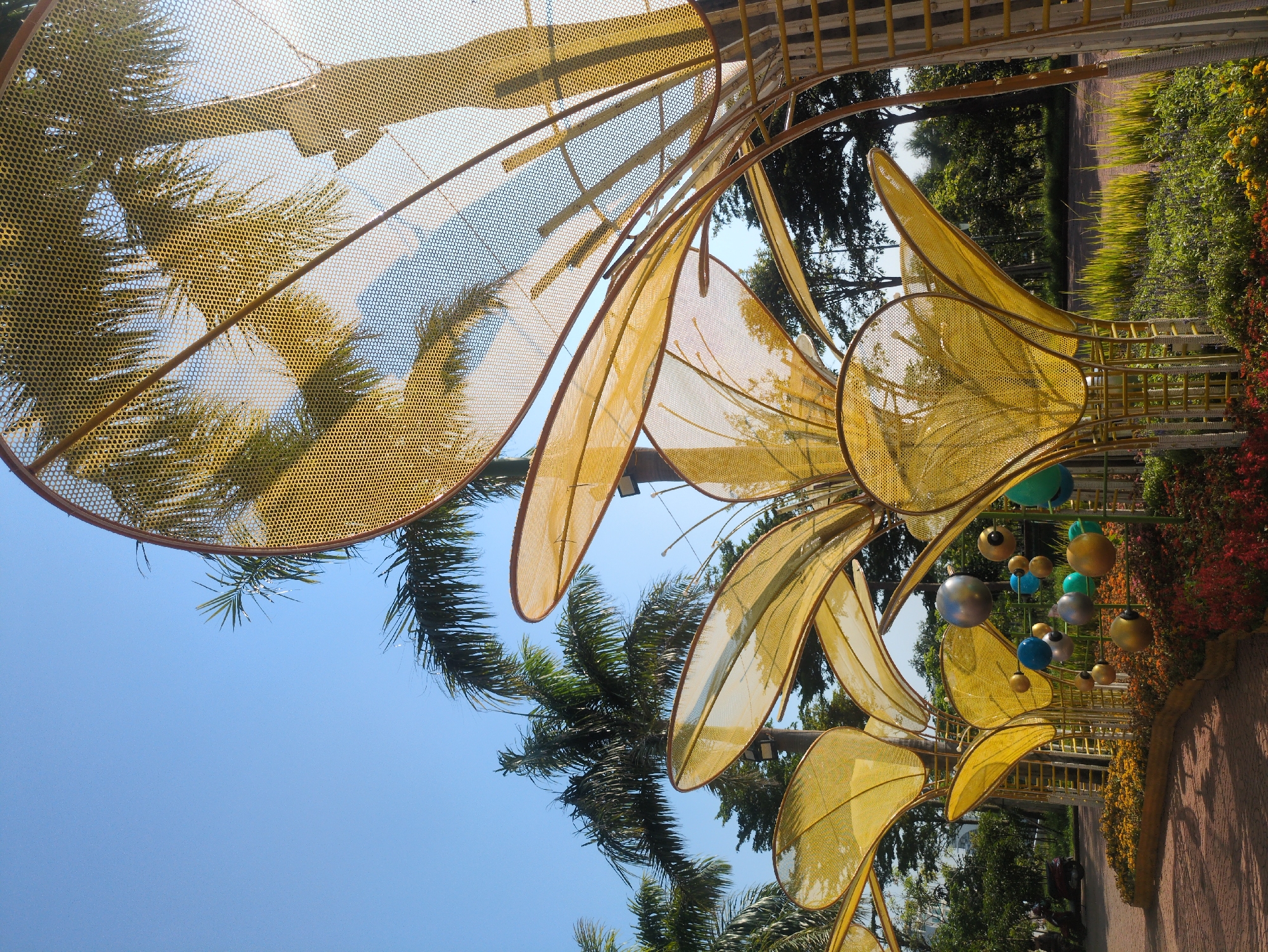 The image presents a tilted view of a public art installation in Tuy Hòa, Vietnam. The installation features large, stylized yellow floral shapes constructed from a mesh material, resembling petals. The structure is partially framed against a clear blue sky and palm trees. To the right, a walkway is lined with vegetation, including flowering shrubs and a variety of plants. Spherical ornaments in colors of blue, green, silver, and gold are suspended from the structure. The scene suggests a sunny day.
