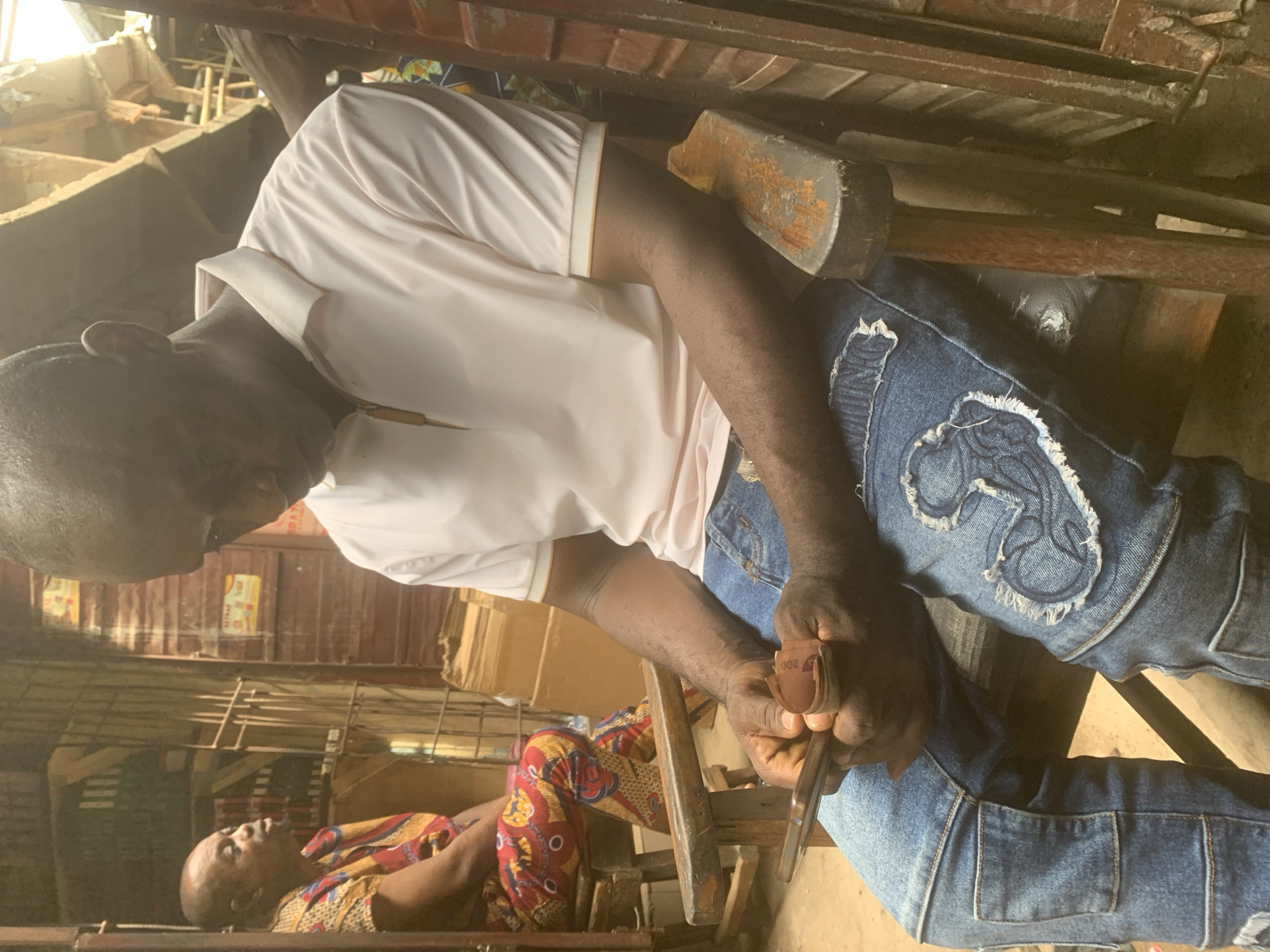 This image, rotated vertically, captures a bustling indoor scene in Fegge, Nigeria, likely a workshop or market stall, under bright daylight.
In the immediate foreground, a dark-skinned man with a shaved head is seated, intently engaged in a task. He wears a light-colored short-sleeved polo shirt with a subtle striped collar and blue denim jeans. A distinctive feature of his attire is the pair of frayed, light-blue patches on his thighs, each intricately cut and sewn to resemble the continent of Africa. His hands are focused on a piece of material, possibly wood or leather, which he is working on with a tool, likely shaping or sanding it. He holds a strip of sandpaper or abrasive material, which clearly shows the number "200" printed on it, indicating its grit.
Behind the working man, slightly out of focus and lower in the frame, another dark-skinned man with a shaved head is visible. He appears to be reclining or lying down on a surface, looking upwards. He is dressed in a brightly patterned shirt, featuring red, yellow, and blue geometric designs, indicative of traditional African fabric.
The surrounding environment consists of visible wooden structures, including beams and shelves, suggesting a functional, possibly rustic, workspace. Cardboard boxes are stacked in the background, hinting at stored goods or materials. The natural light illuminates the scene, creating a sense of activity and diligence within this localized setting.