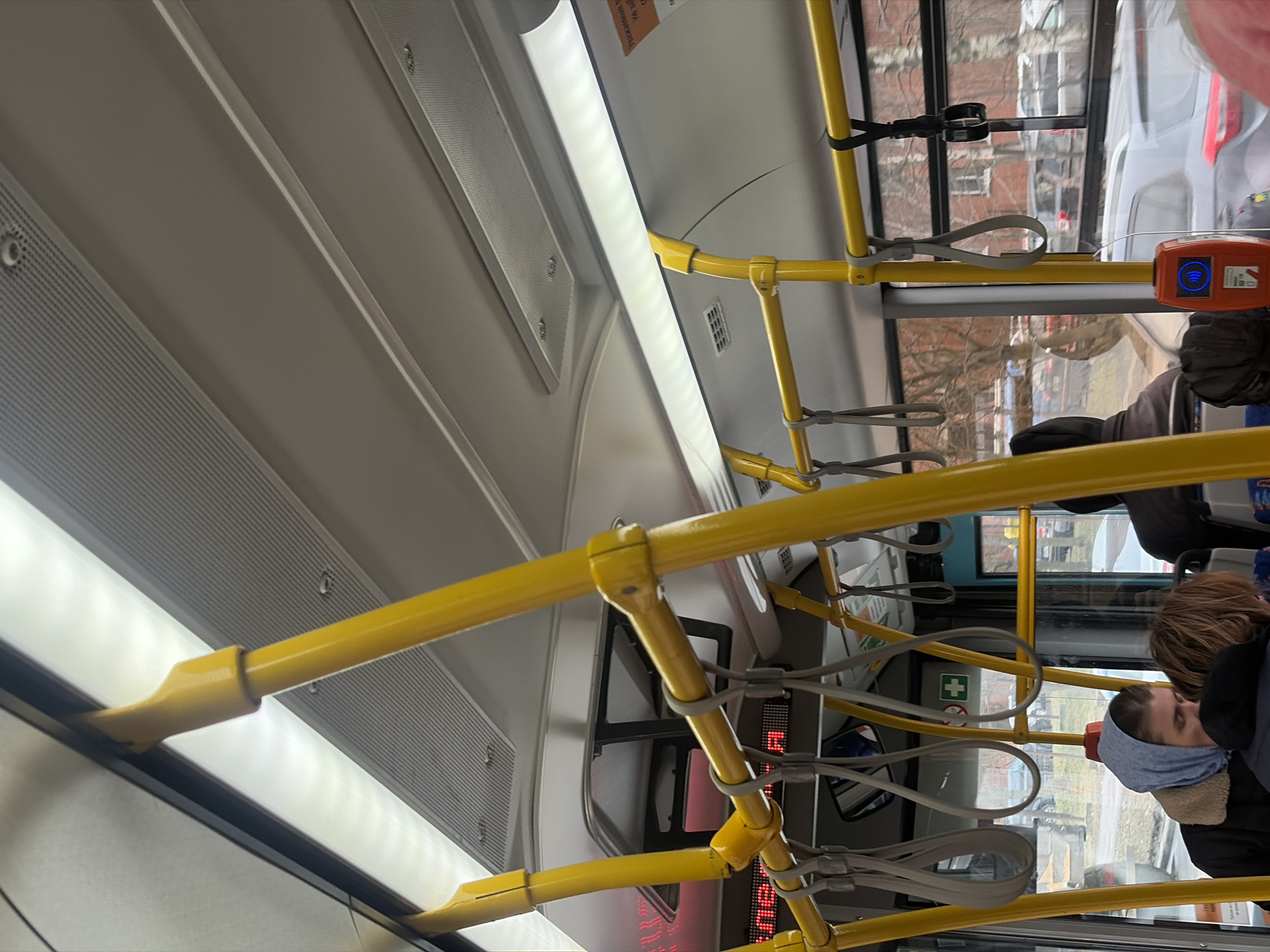 The image depicts the interior of a public transport vehicle, likely a bus or tram, in what appears to be Saint Petersburg, Russia, based on the architecture visible through the windows. The viewpoint is looking upwards towards the ceiling, which features lighting panels and ventilation grilles. Yellow handrails and straps are present, providing support for standing passengers. Through the windows, a cityscape is visible, with buildings and trees. Several passengers are present, including one with a blue head covering and another with brown hair. A ticket validation device is affixed to the handrail. A first aid kit sign is attached to the wall.