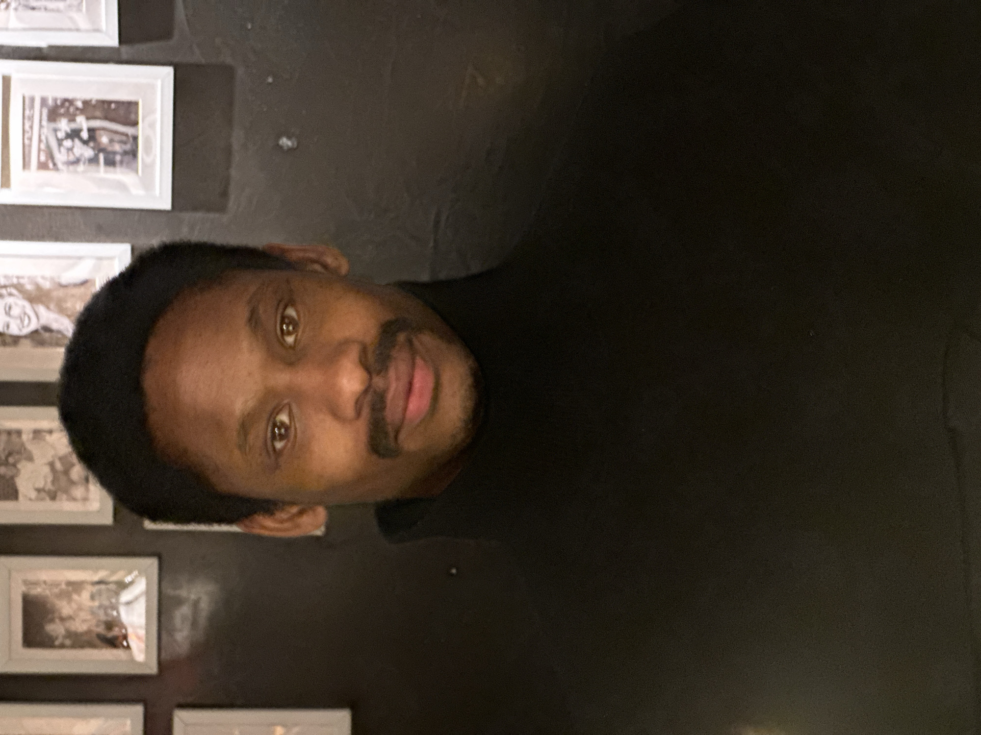 An adult male with dark skin, short dark hair, dark eyes, and a mustache is centrally positioned, looking directly at the viewer with a neutral expression. He is wearing a black turtleneck sweater. The background consists of a dark wall displaying multiple light-framed pictures. One frame to the upper right of the subject's head depicts a light-haired female. Another frame to the right contains text, including the word "FLAGSTAFF". This detail indicates the location is likely the Flagstaff restaurant, situated at 238 King's Road, London SW3 5UE, United Kingdom. The scene is illuminated primarily from the front, highlighting the subject.