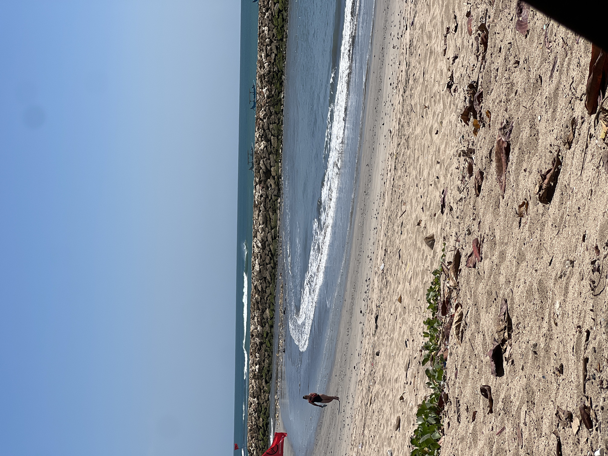 An aerial view of a beach in Kuta, Indonesia, shows a person walking near the shoreline. The beach is sandy and scattered with debris, including leaves and rocks. In the distance, waves crash against a stone jetty, and several boats are visible on the ocean. The sky is clear and blue.