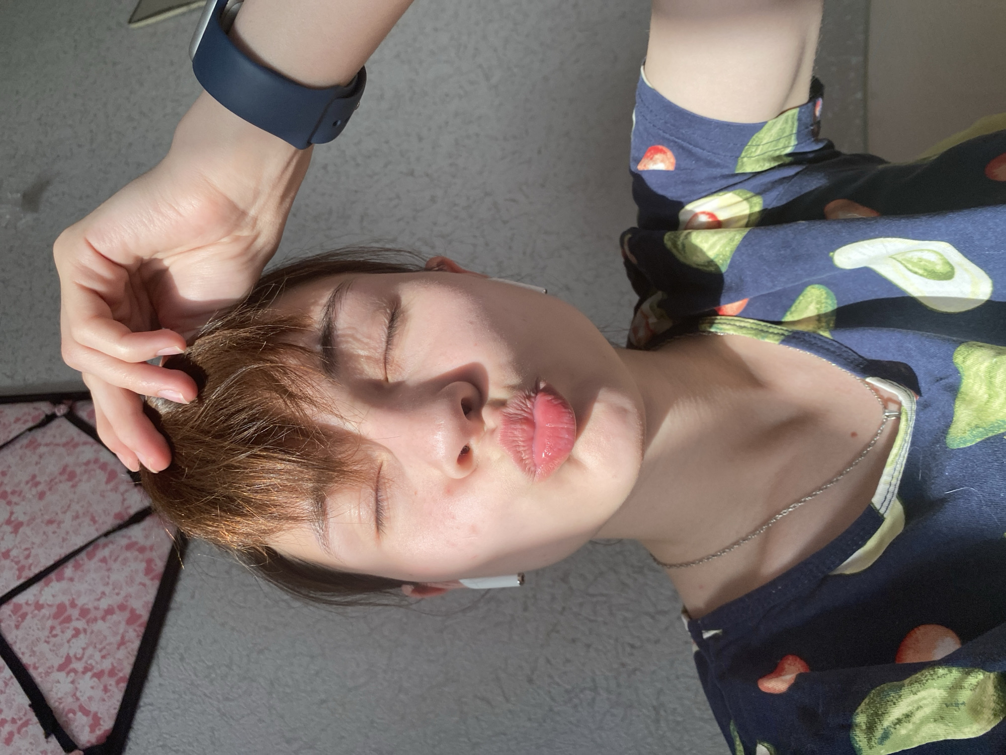 A person with closed eyes and pursed lips is lying down. The person is wearing a dark blue shirt with an avocado and tomato print, and a silver chain necklace. The person's right hand is resting on their head and they are wearing a blue smartwatch. The person has short brown hair with light brown highlights. The person is lying on a gray surface. A patterned object with pink and black triangles is partially visible on the left side of the image. The location is not specified in the image. Therefore, the location of the city of Votkinsk, Russia is not supported by the image.