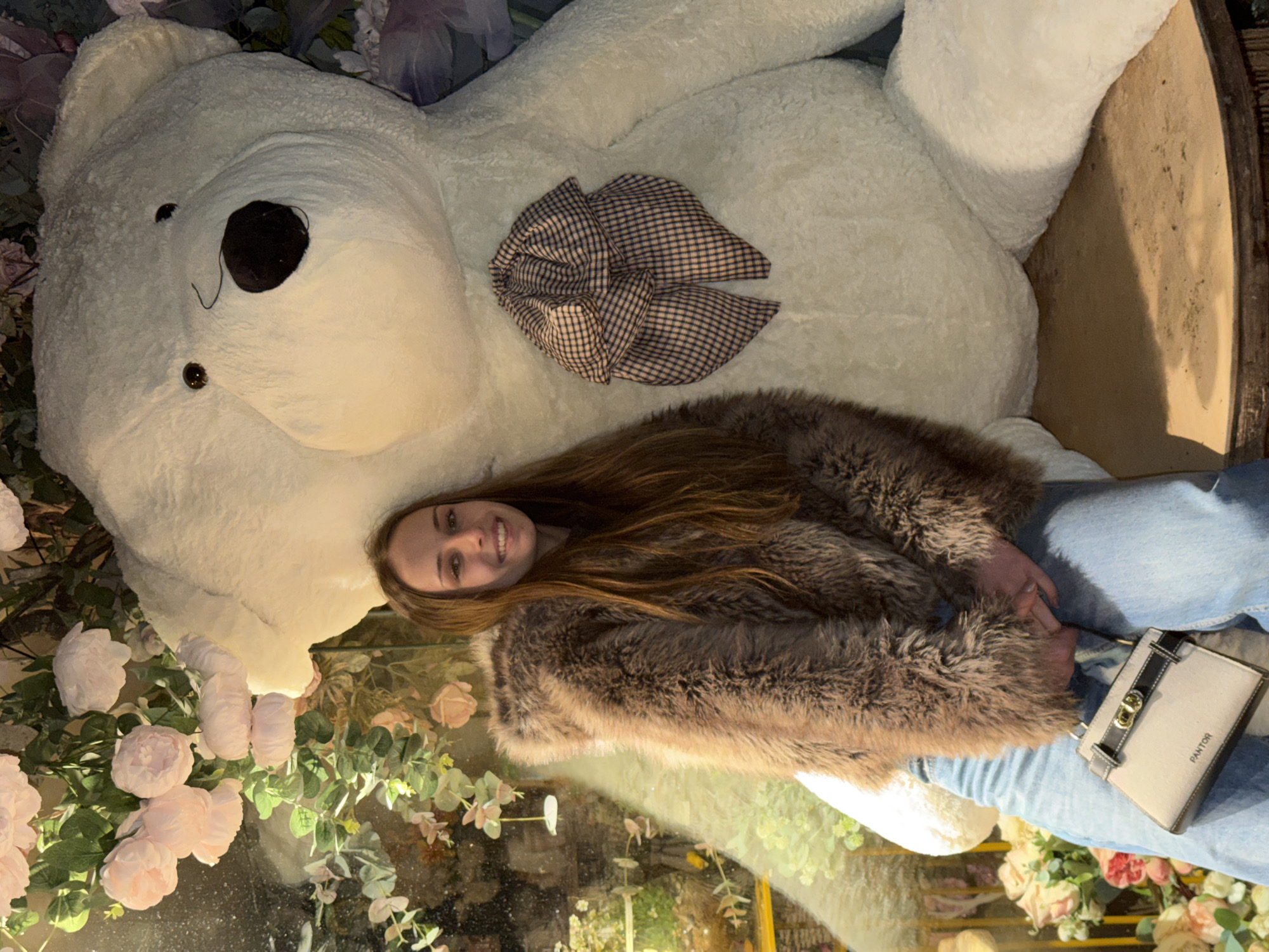 A young woman lies on a large white stuffed bear, likely for a photograph, amidst an abundance of artificial flowers and foliage. The woman is wearing a fur coat and jeans, and she is holding a white and black handbag labeled “PANTON”. The bear has a gingham bow on its chest. The setting seems to be an indoor space designed for photo opportunities, possibly a shop or studio in Sofia, Bulgaria, given the request. The image appears to be taken from an overhead perspective.
