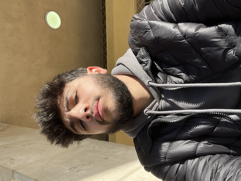 A young man with dark hair and a trimmed beard looks at the camera. He's wearing a grey hooded sweatshirt under a puffy black jacket. The light illuminates his face, highlighting his features. The background appears to be an exterior wall with a round, recessed light. The man is slightly tilted to the left in the frame.