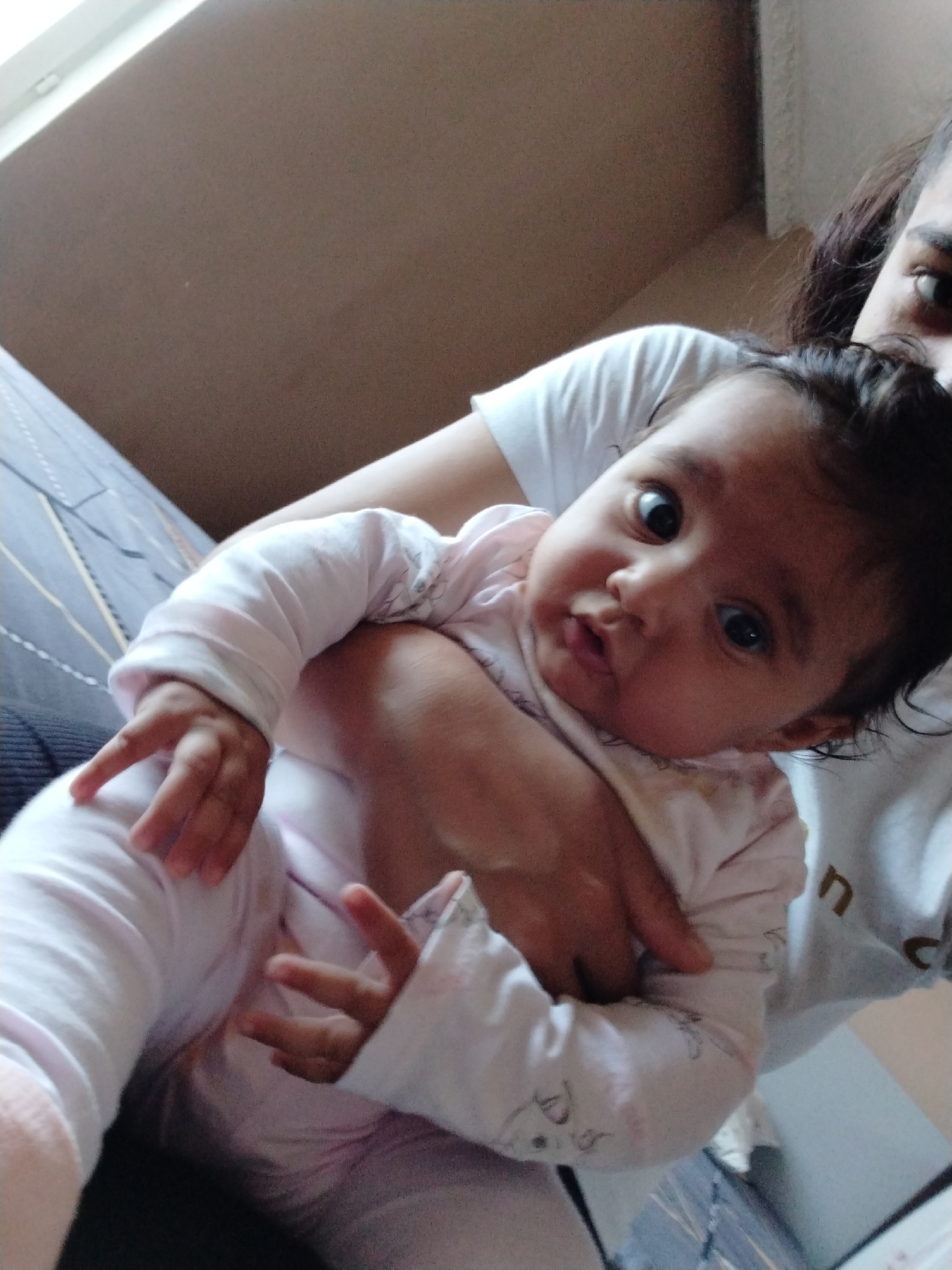 A baby wearing a white patterned shirt is being held by a person wearing a white shirt. The baby is looking at the camera with wide eyes. The background is a light brown wall, and part of a bed with a striped cover is visible. The location and city are not specified in the image.
