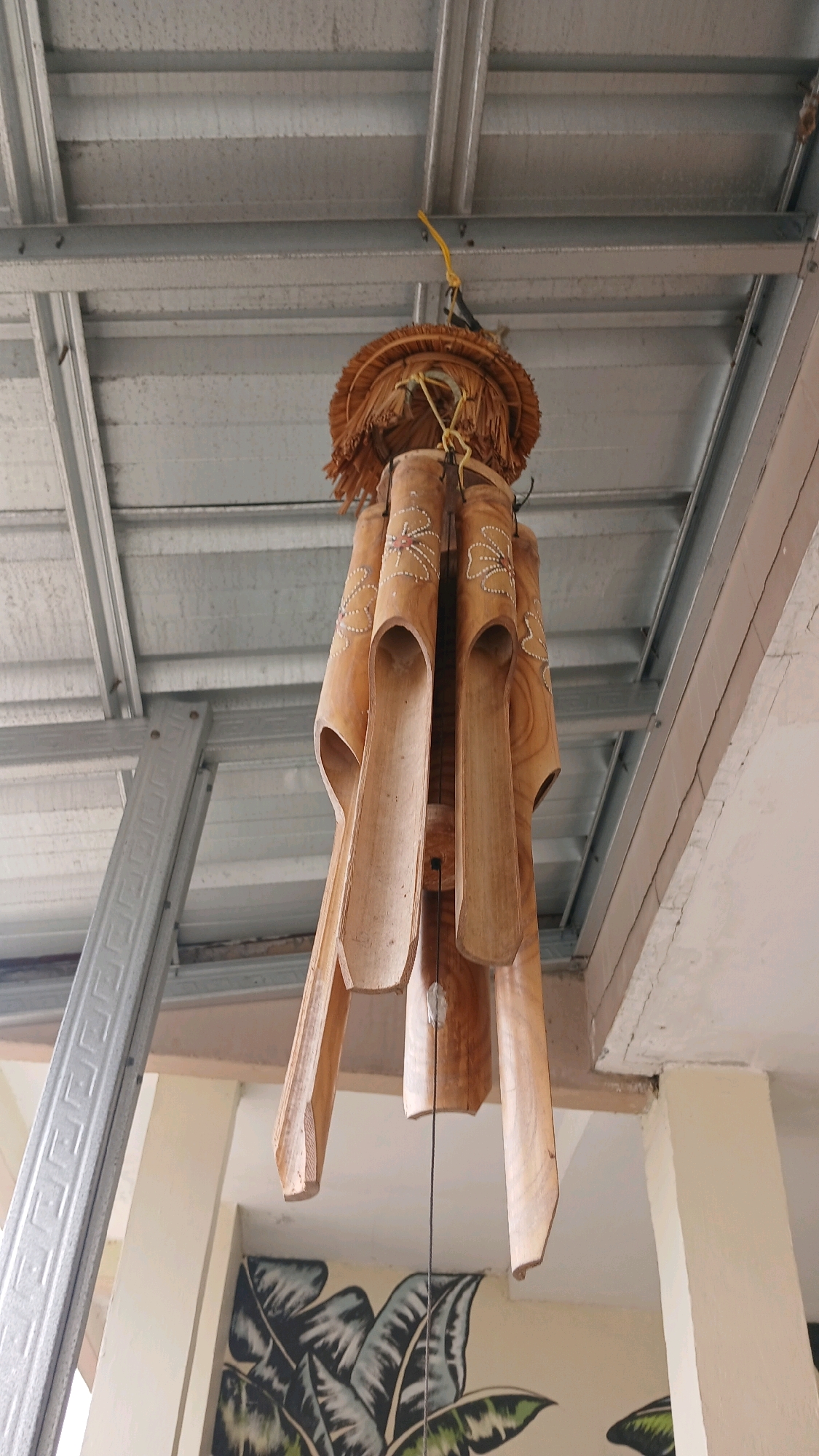 A wooden wind chime made of hollow bamboo tubes hangs from a metallic ceiling structure. The wind chime is adorned with carved floral patterns. Below the wind chime, a wall features a mural of large, dark green leaves. The scene is indoors, likely in a covered outdoor area, given the exposed ceiling beams and a visible pillar. The location is specified as Melatiwangi, Indonesia.