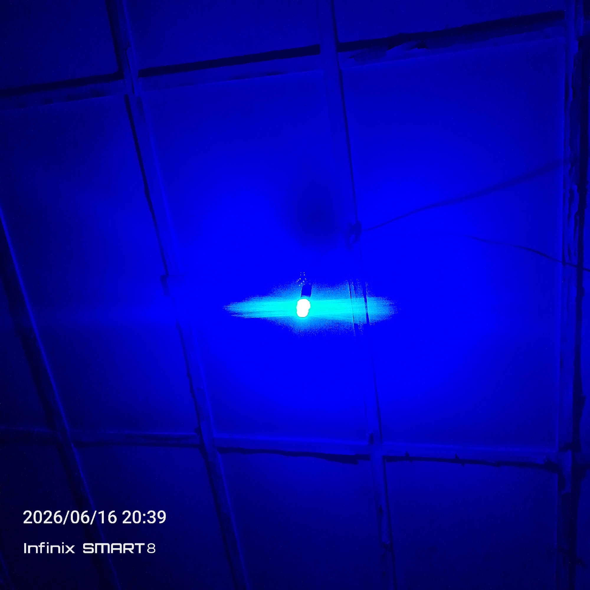 The image shows a close-up, low-angle shot of a ceiling with a blue light bulb illuminated in the center. The ceiling appears to be made of panels supported by wooden beams, creating a grid-like structure. The blue light casts a strong glow, illuminating the immediate area around the bulb and casting faint shadows on the ceiling panels. The overall color palette is dominated by deep blues, with the bright white light of the bulb being the focal point. The timestamp "2026/06/16 20:39" and device information "Infinix SMART 8" are overlaid in white text on the bottom left of the image. There are no people or discernible actions visible. The environment is dark and dimly lit, with the primary light source being the blue bulb. Based on the information provided, this image is from Jos, Nigeria.