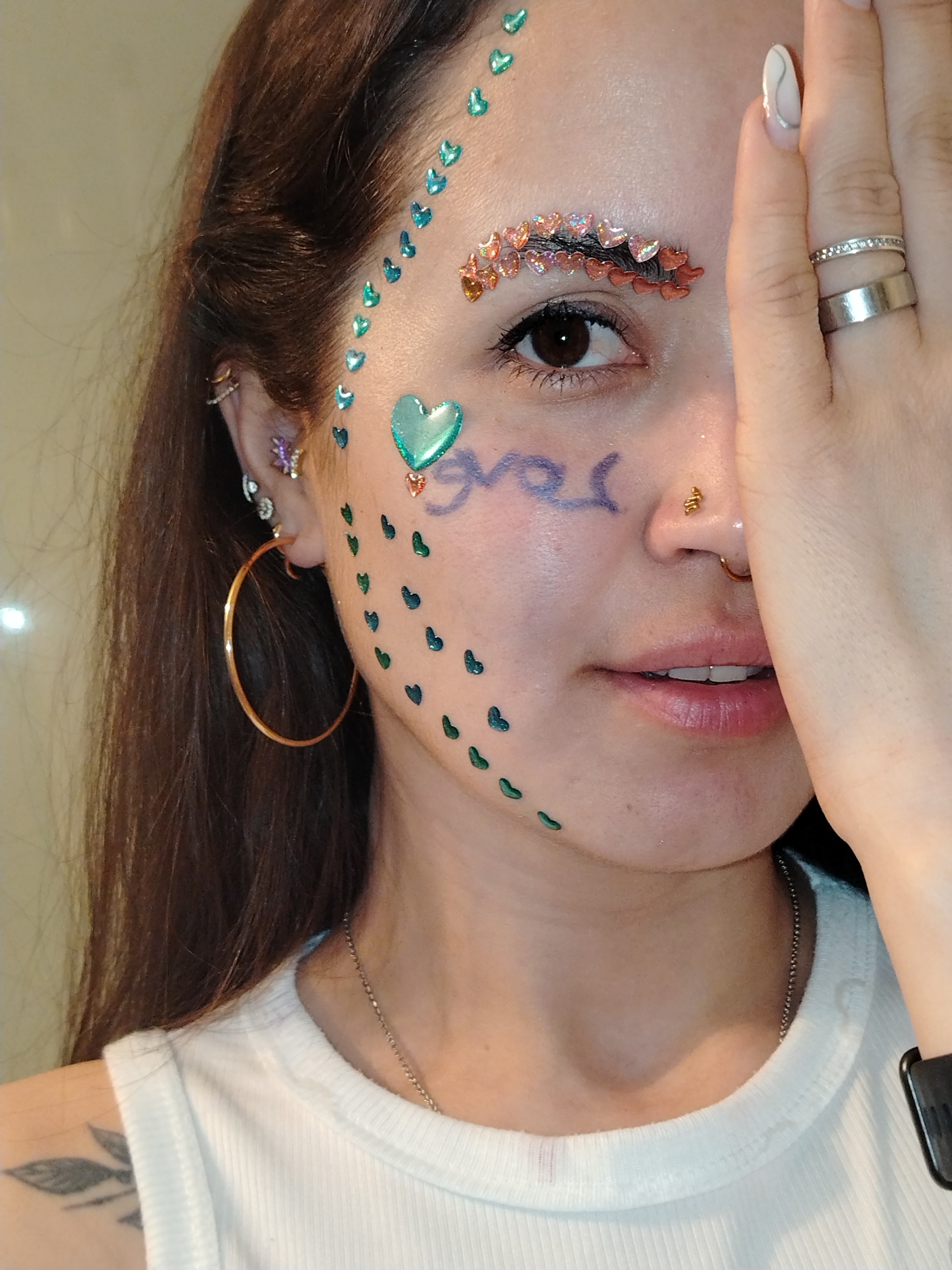A woman is visible from the chest up, facing the camera, with her hand partially obscuring her face. She has dark hair and fair skin. Her face is adorned with colorful stickers in the shape of hearts and other embellishments. One eyebrow is completely covered with small, red and orange heart-shaped stickers. A large, green heart sticker is on her cheek near the corner of her eye, with smaller green heart stickers cascading down her face. There is a blue scribbled word, possibly in Russian or another Cyrillic alphabet, near her nose. She has several piercings: one in her nostril, one in her septum, and multiple in her left ear, including a large gold hoop earring and smaller decorative earrings. She is wearing a white ribbed tank top and a thin silver necklace. A dark tattoo is visible on her left shoulder. She is wearing two silver rings on her ring finger. The background is indistinct and appears to be a light-colored wall. There is nothing to suggest that the image was taken in Noviy, Russia.