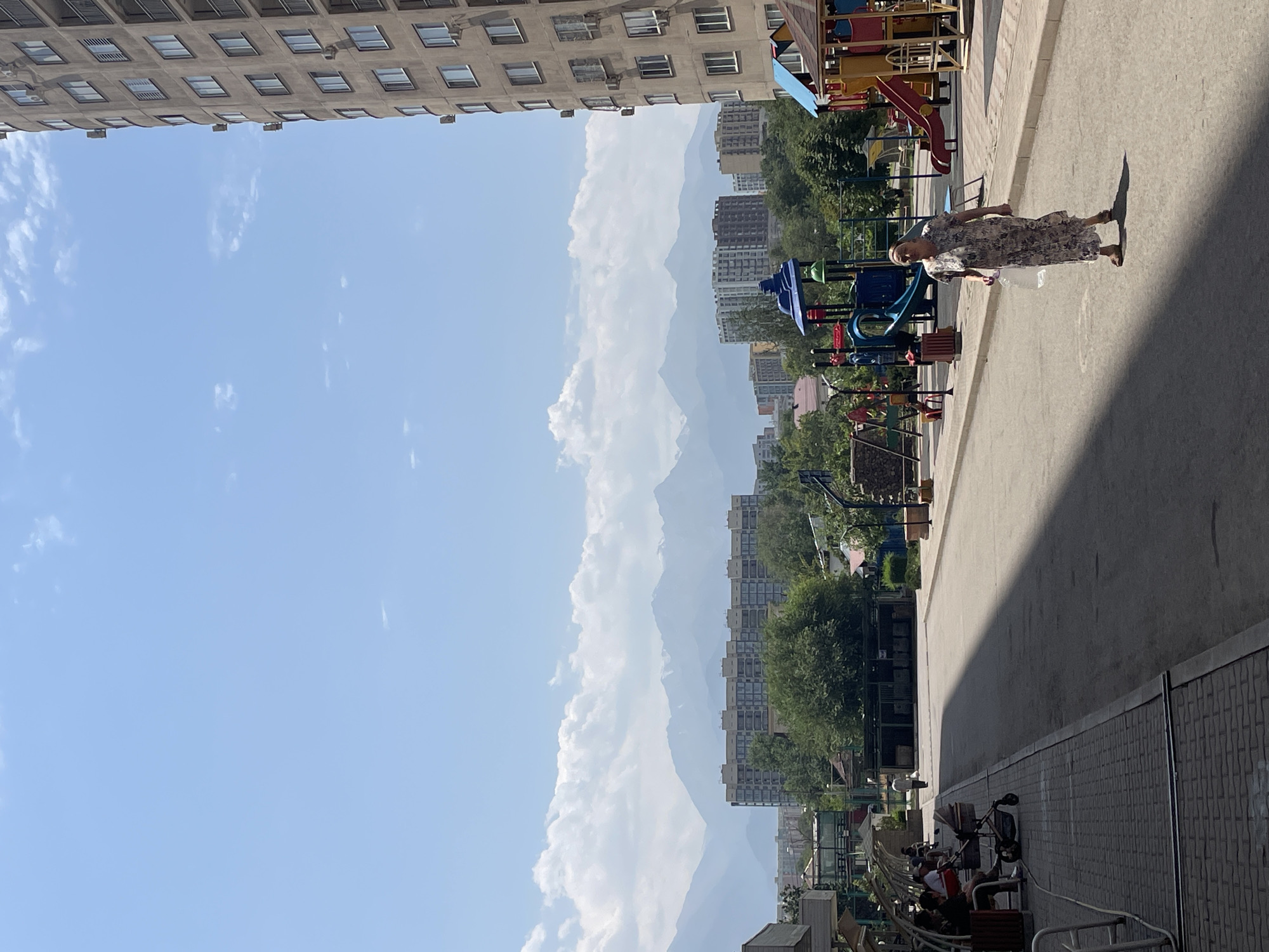 The image depicts a sunny day in Almaty, Kazakhstan, with a clear blue sky dotted with sparse white clouds. In the foreground, a paved walkway runs alongside a residential street, casting a long shadow across the pavement. On the left side of the walkway, several people are seated on benches, some with children and strollers. To the right, a colorful playground with slides and swings is visible, with a woman walking towards the viewer. In the background, apartment buildings and lush green trees frame the view of majestic, snow-capped mountains, a characteristic feature of Almaty's landscape. The overall scene suggests a peaceful residential area with recreational facilities, set against a backdrop of natural beauty.