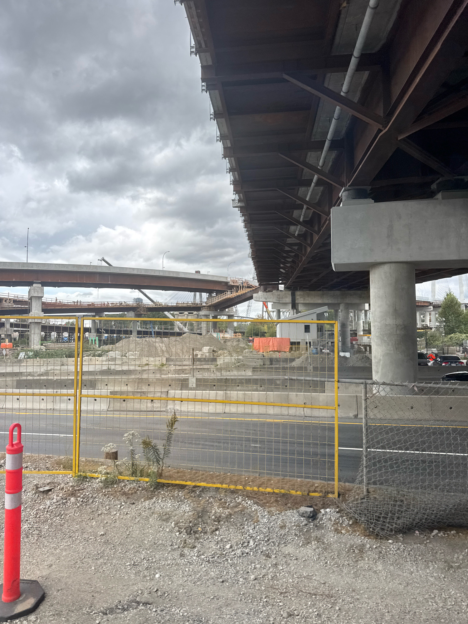 The image depicts an under-construction overpass in Surrey, Canada, under a cloudy sky. A large, multi-tiered concrete and steel overpass dominates the scene, with visible structural beams, pipes, and supporting pillars. Construction materials such as piles of sand and concrete barriers are present in the mid-ground, along with temporary structures like a small white building and an orange tarp. A yellow chain-link fence with a bright yellow top rail separates the construction area from a paved road, where a red traffic cone with reflective grey bands is positioned on the left. In the background, other elevated road structures and distant buildings are visible. The overall environment is industrial and appears to be a busy construction site.