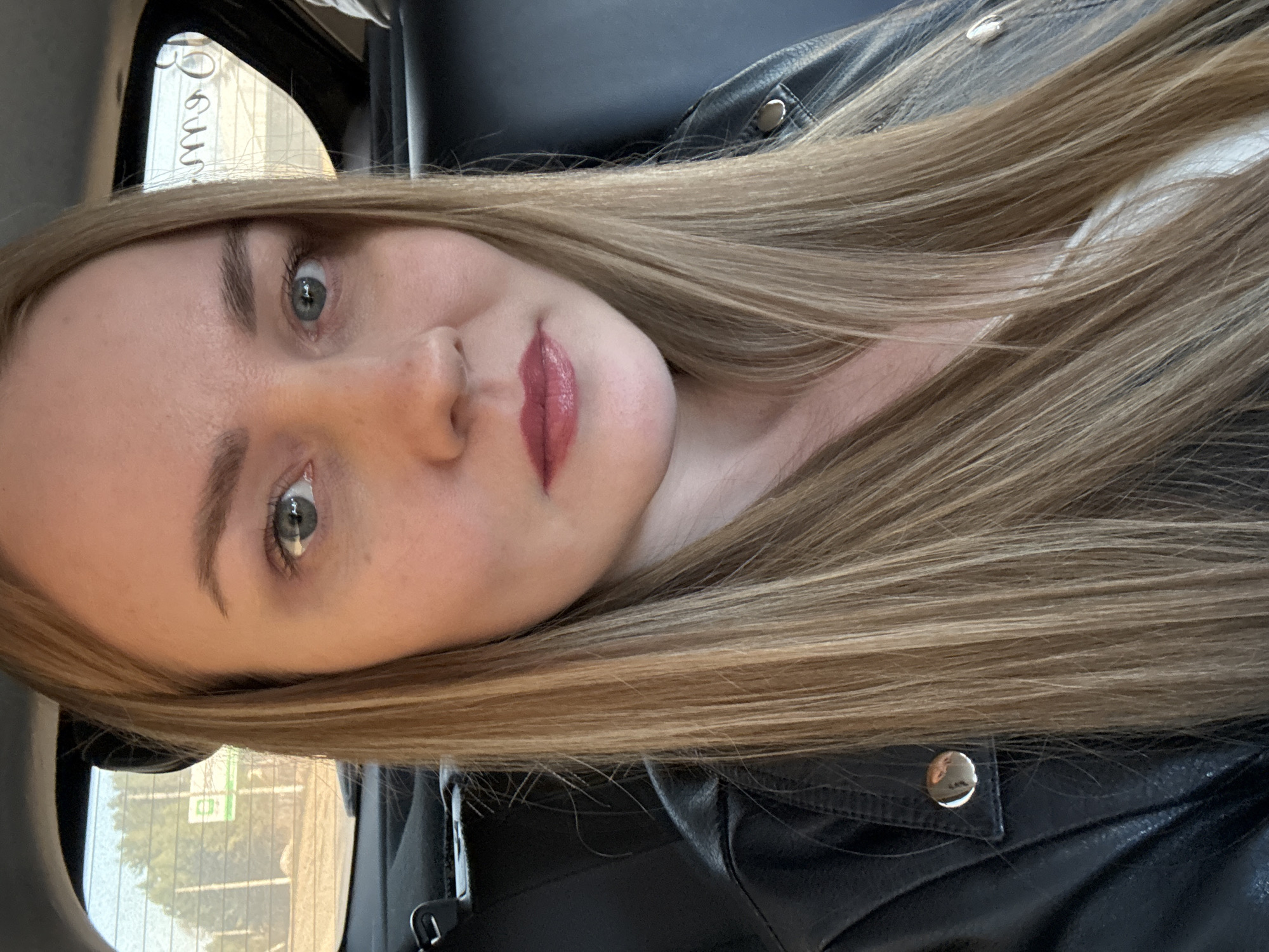 A young woman with blonde hair and fair skin is looking at the camera. She is wearing a black leather jacket. The background suggests she is in a vehicle, possibly a car. Through the window, there is a blurred view of a city street in Chaikovsky, Russia, with buildings and trees visible.